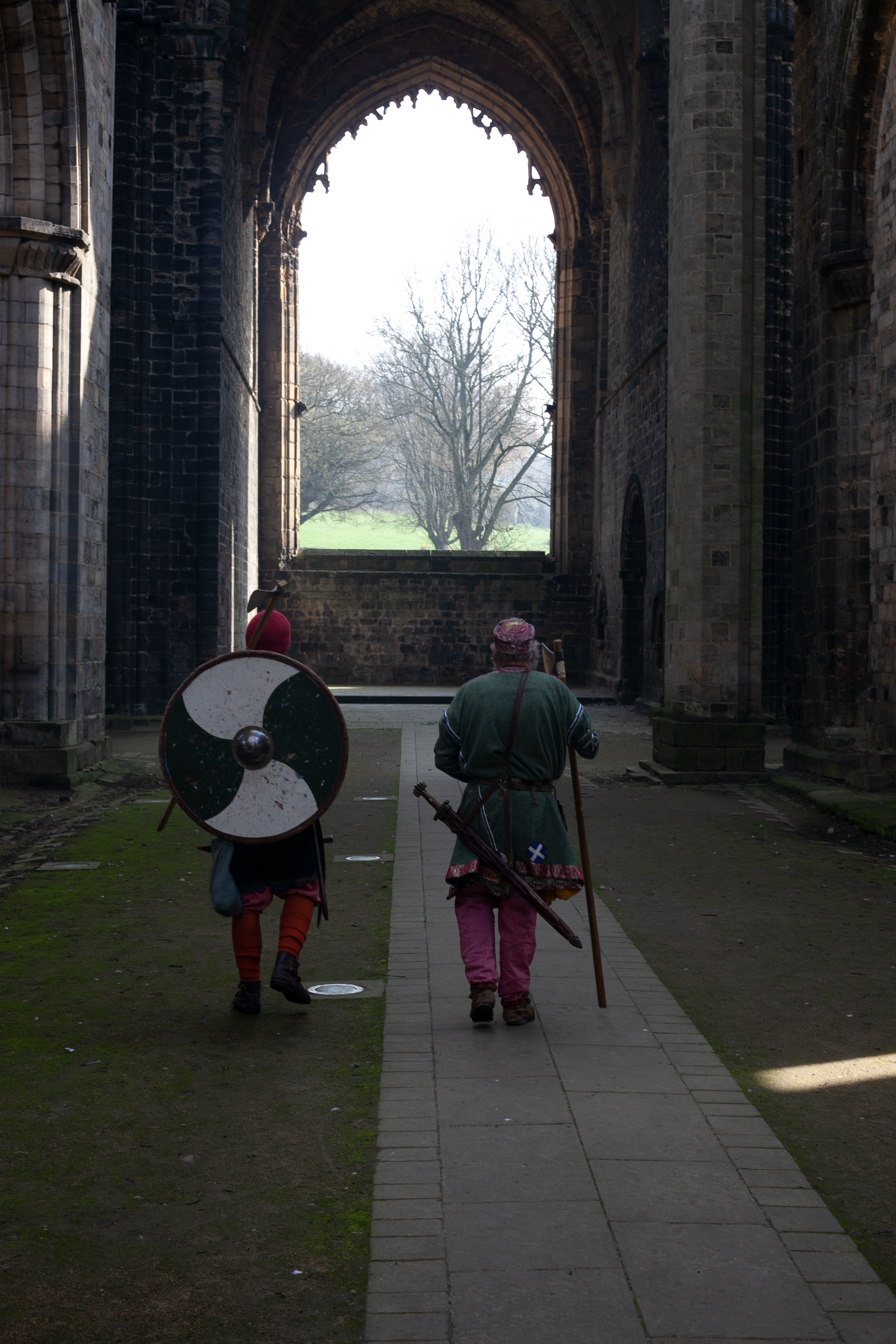 Two people walking through an old stone archway, dressed in medieval-style clothing, one carrying a round shield and the other with a sword at his side, with a grassy area and trees visible outside.