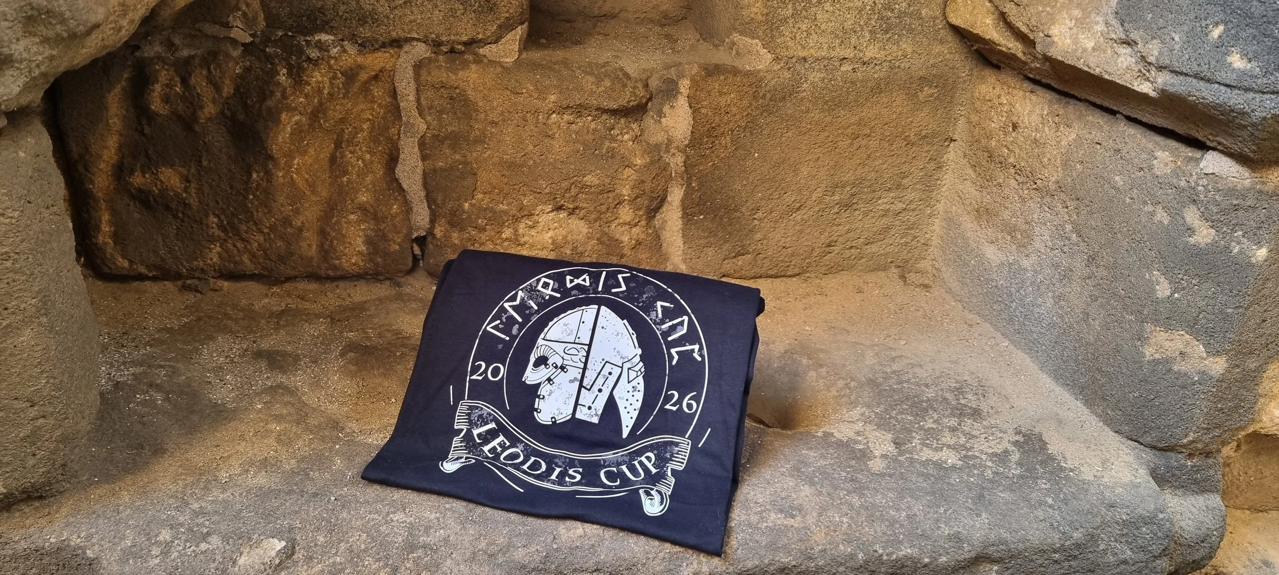 T-shirt with Leodis Cup logo, featuring a stylized image of Viking and medieval helmet, placed on a stone surface in front of a stone wall.