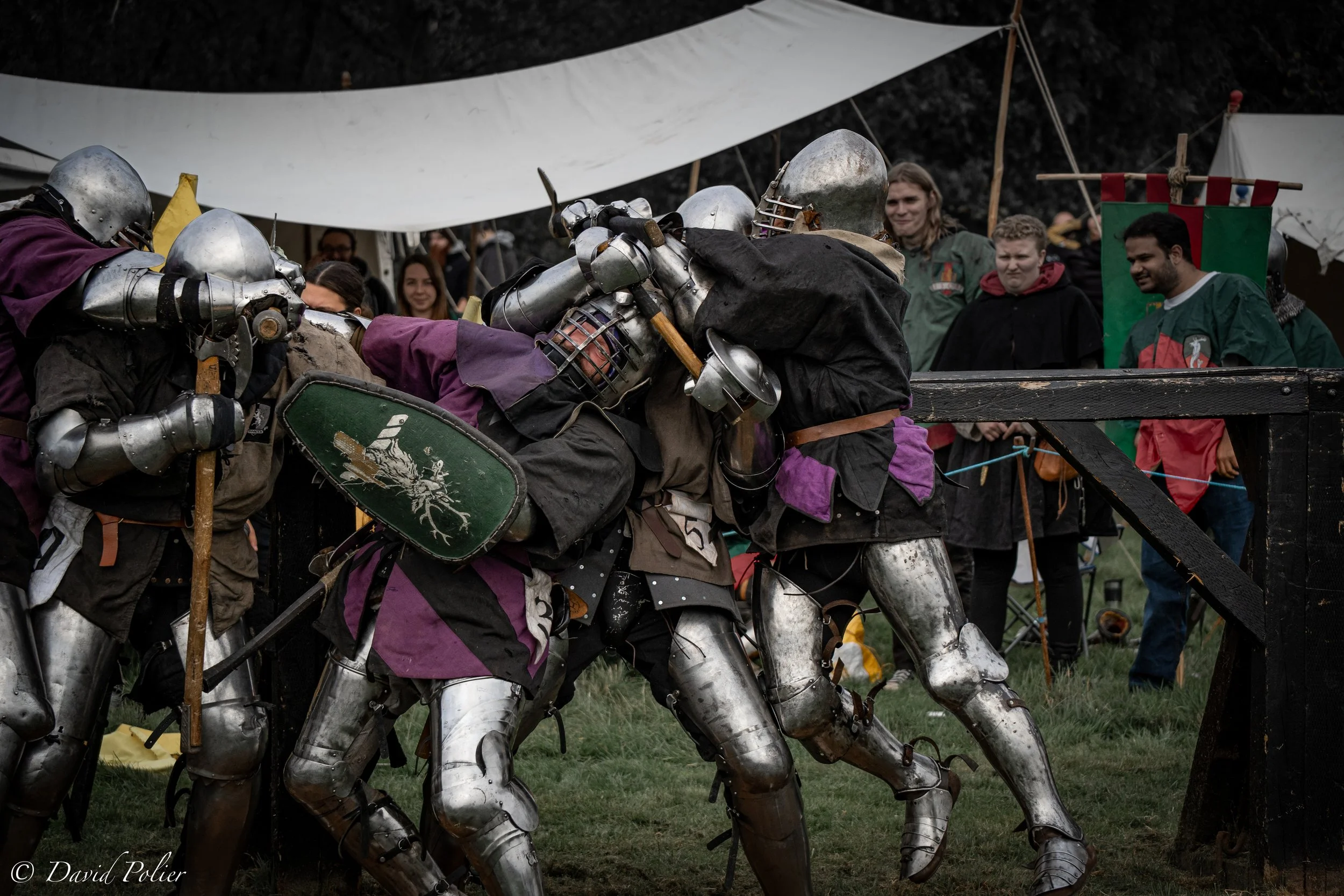Medieval reenactors dressed as knights in armor battling with wooden shields during a jousting event, with a crowd of spectators in medieval-themed costumes watching in the background.