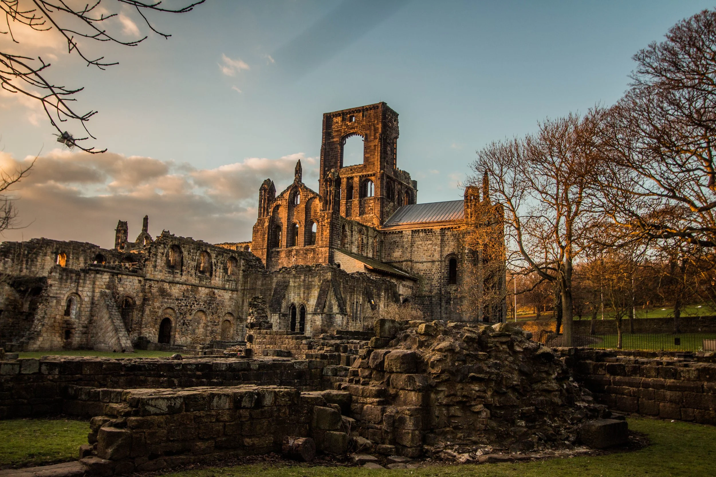 Ruins of an old stone church or castle at sunset, with leafless trees around and a partly cloudy sky in the background.
