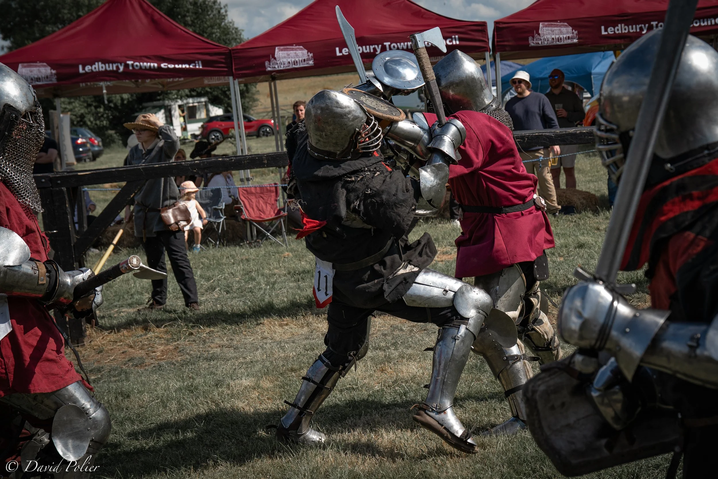 Two people dressed in medieval armor engaging in full contact martial art fighting competition during a reenactment event, with spectators and red tents in the background.