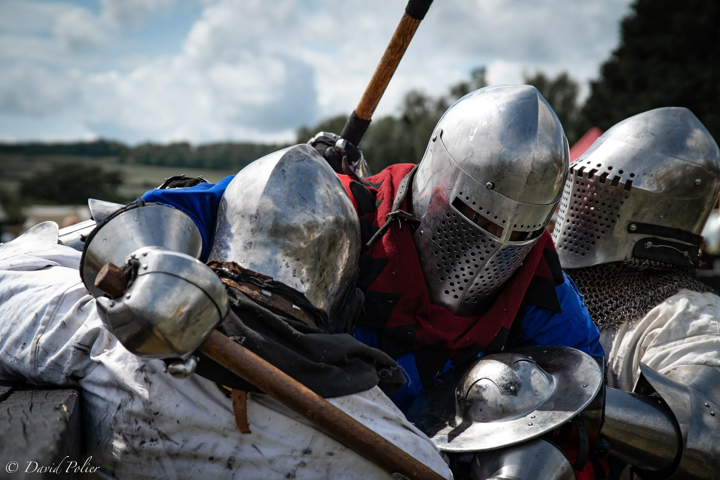 Two medieval knights in shiny armor fighting in full-contact martial art fighting competition event outdoors, with a cloudy sky and green landscape in the background.