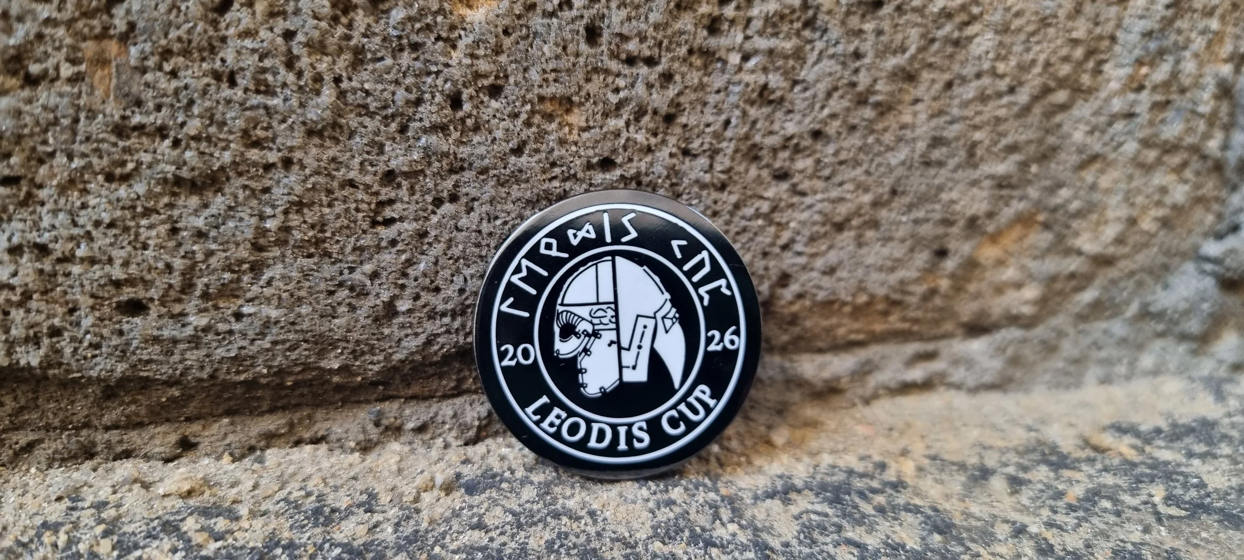 A round badge with a black background and white graphics, featuring a medieval knight's helmet and a Viking helmet, with the text "LEODIS CUP" and the year "2026" on it, placed against a rough stone surface.