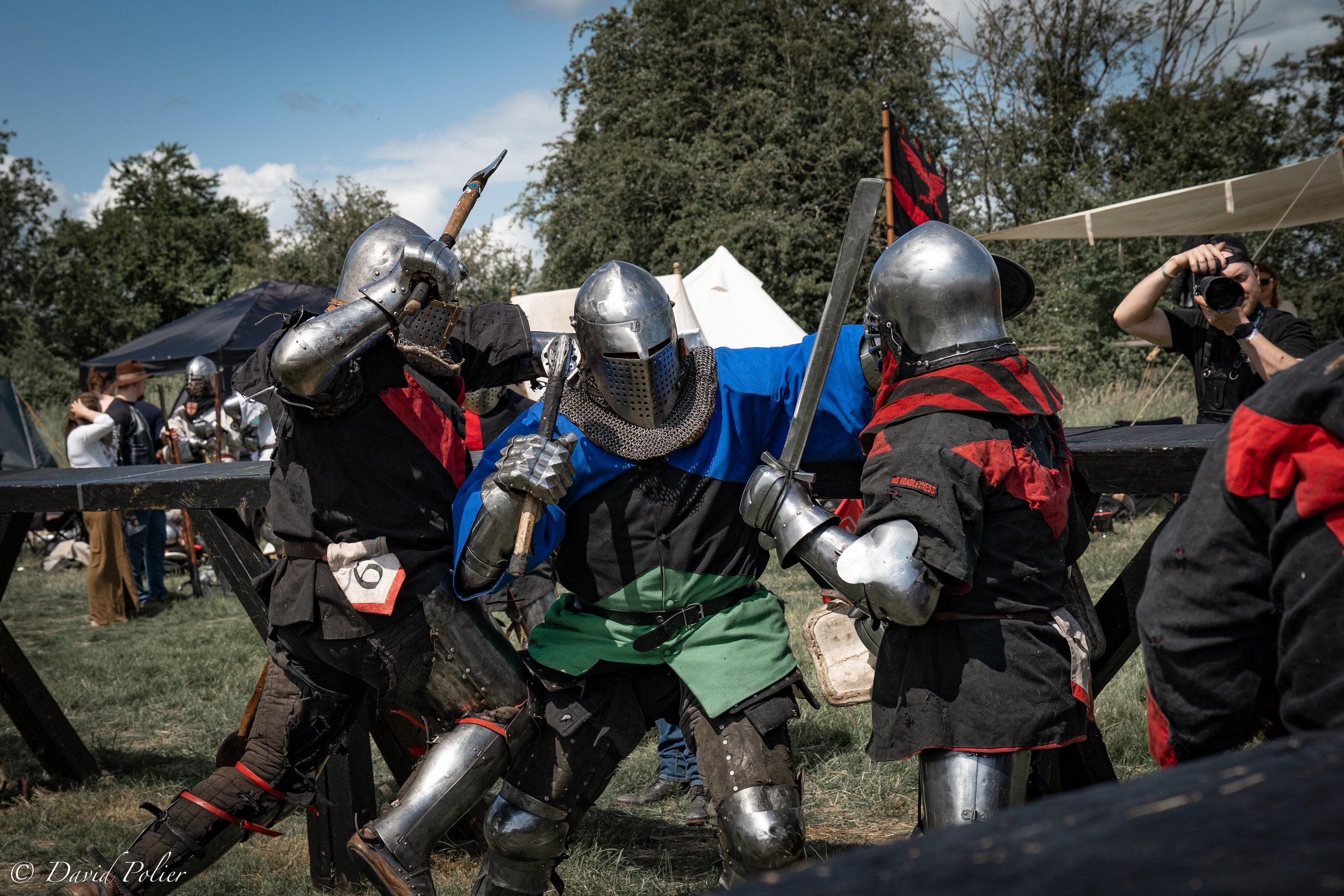 People dressed in medieval armor engaging in full contact historical martial art fighting competition outdoors.