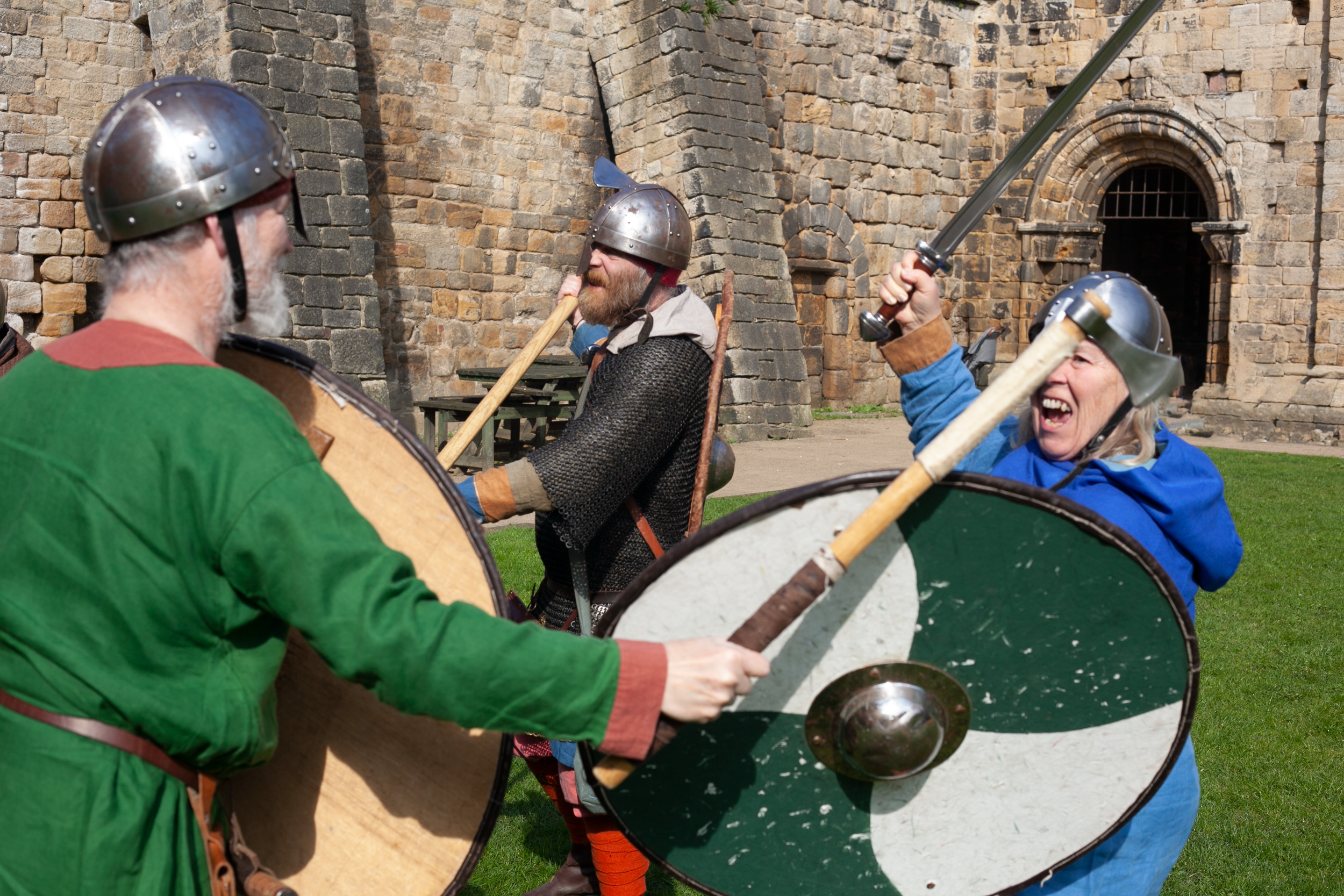 Three people participating in a reenactment or fair dressed as viking warriors with helmets, shields, and swords outside a stone castle