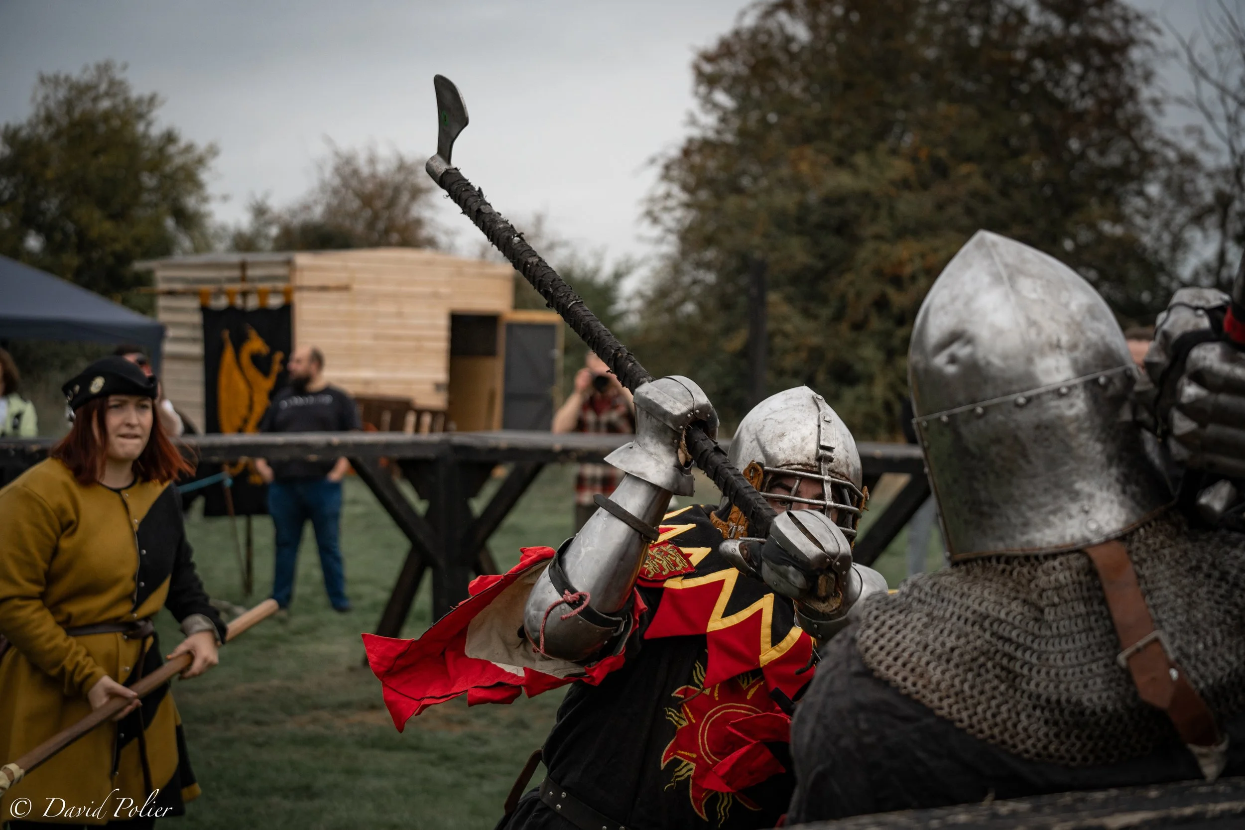 Two medieval knights in armor fighting with swords during full-contact martial art fighting competition, with spectators and a wooden structure in the background.