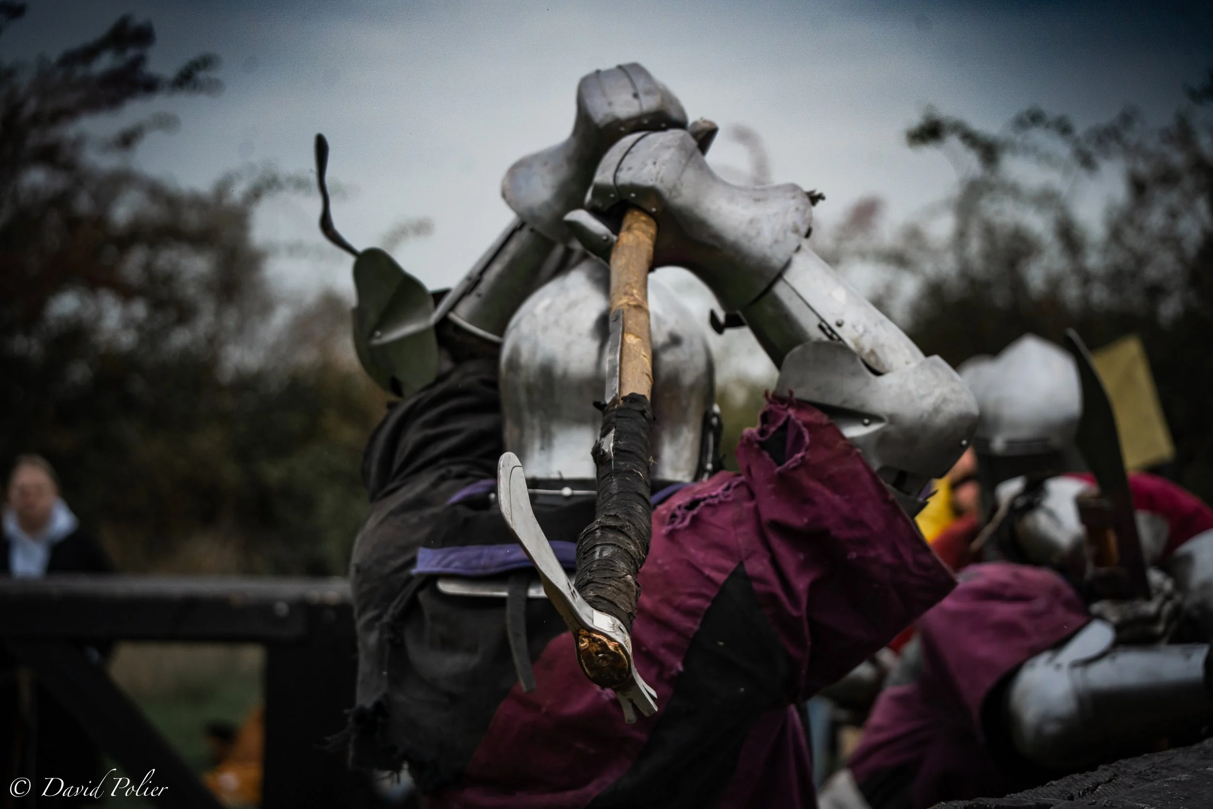 People dressed as medieval knights in armor engaged in a reenactment or battle, outdoors on a cloudy day.