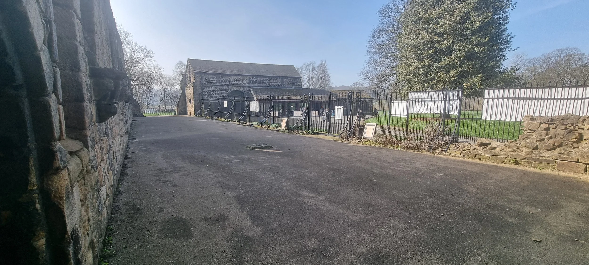 A paved pathway runs along a stone wall on the left and a black metal fence on the right, with a historic stone building and trees in the background on a clear day.
