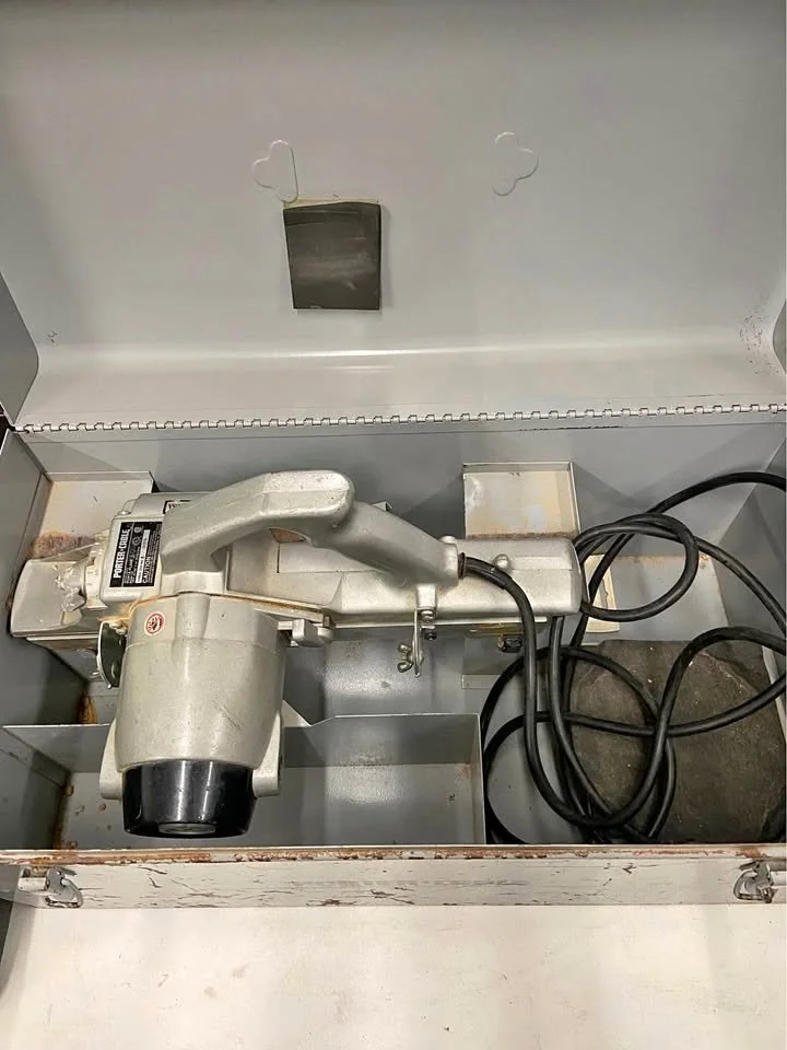 An old metal meat grinder inside a metal storage box with some black electrical cords and a small metal container.