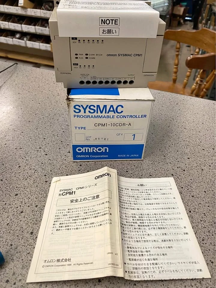 An Omron SYSMAC programmable controller and its packaging box on a speckled gray countertop, with instruction manuals underneath, in a hardware store with shelves of small parts in the background.