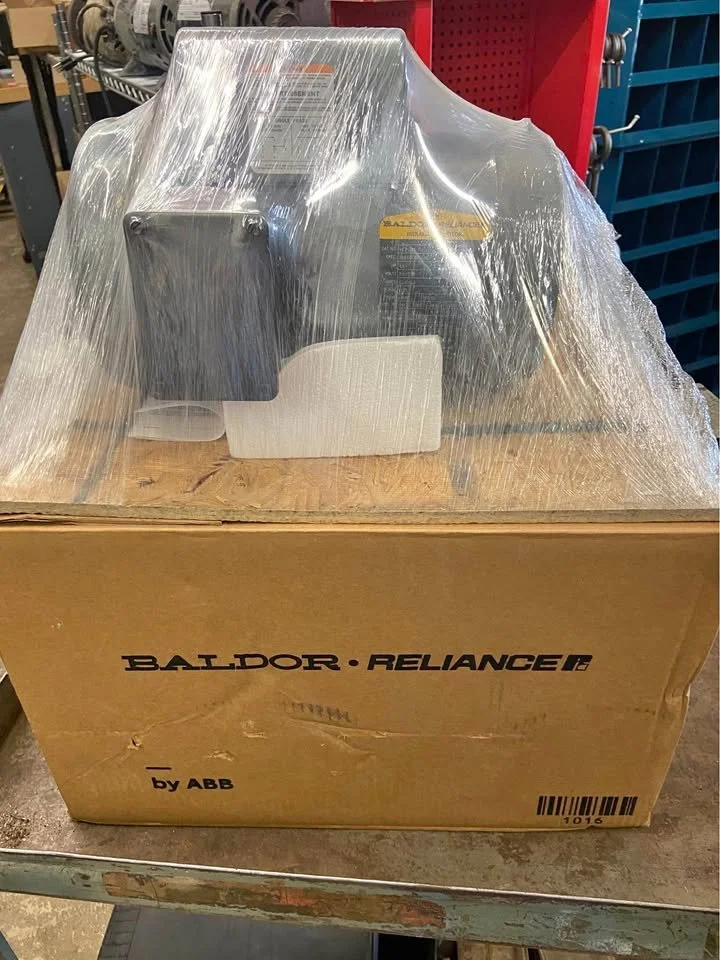 A large industrial machine on a wooden pallet, wrapped in clear plastic, on top of a cardboard box labeled 'BALDOR RELIANCE by ABB' in a warehouse setting.