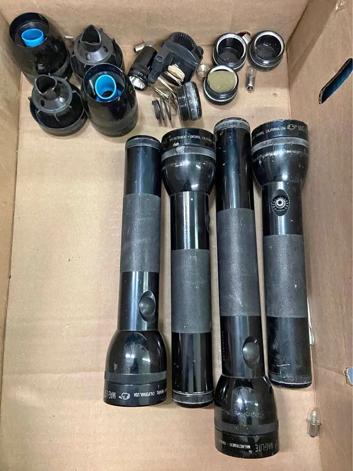 Four black telescopic flashlights with textured grips inside a cardboard box, along with various camera lens adapters and accessories at the top.