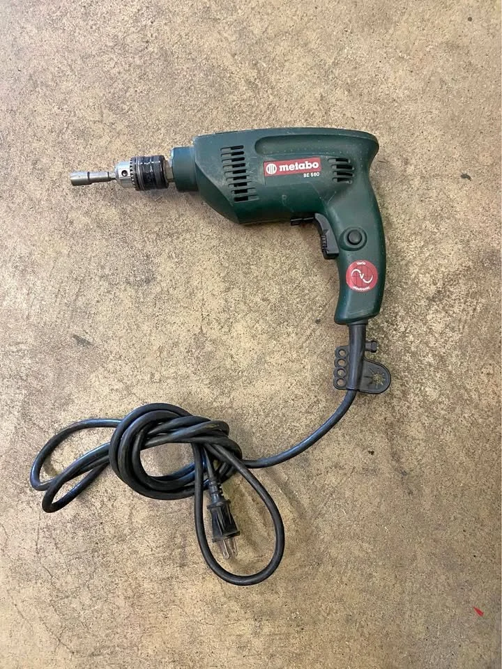 A green Metabo electric drill with a cord and plug, placed on a concrete surface.