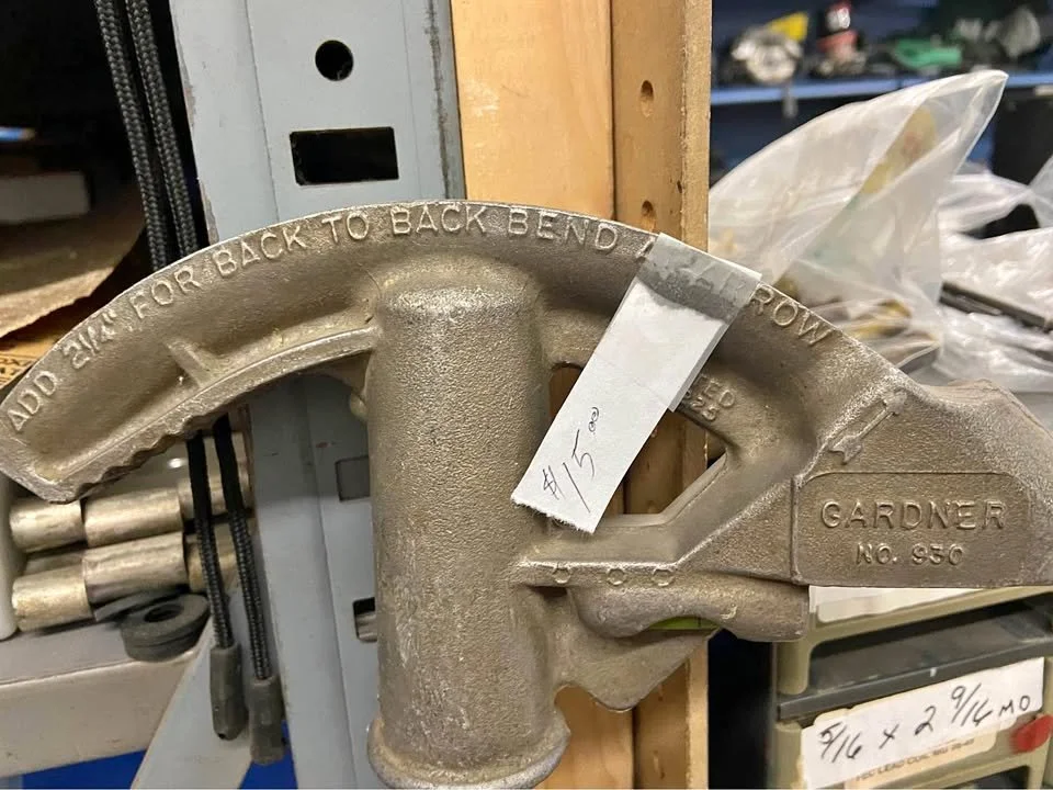 A large Gardner pipe vise with a handle for turning, mounted on a metal frame, with a price tag of $15.00 on it, located in a workshop or hardware store.
