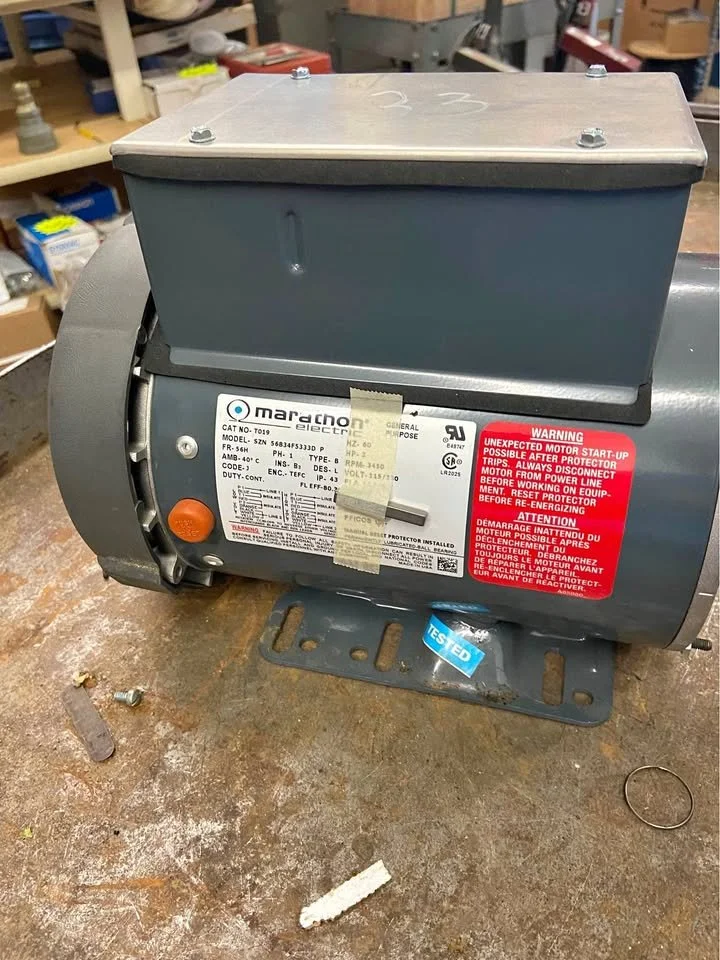 An electric motor with a gray metal casing and a gray metal cover on top, mounted on a metal base plate, situated on a workshop floor with tools and shelves in the background.