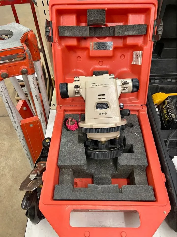 A theodolite or surveying instrument inside a red carrying case with foam padding, placed on a workbench in a workshop or hardware store.