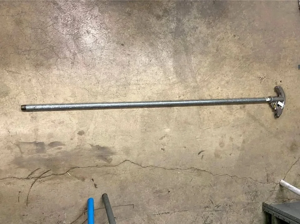 A long metal rod with a rotating wheel attached to one end, lying on a concrete floor.