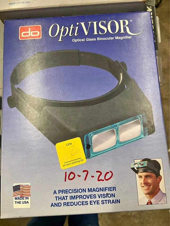 Boxed OptiVISOR optical glass binocular magnifier with headband, label indicates it is made in USA, featuring a picture of a man wearing the magnifier, and a yellow sticker with the number 2 and a note to check it on the packing list.