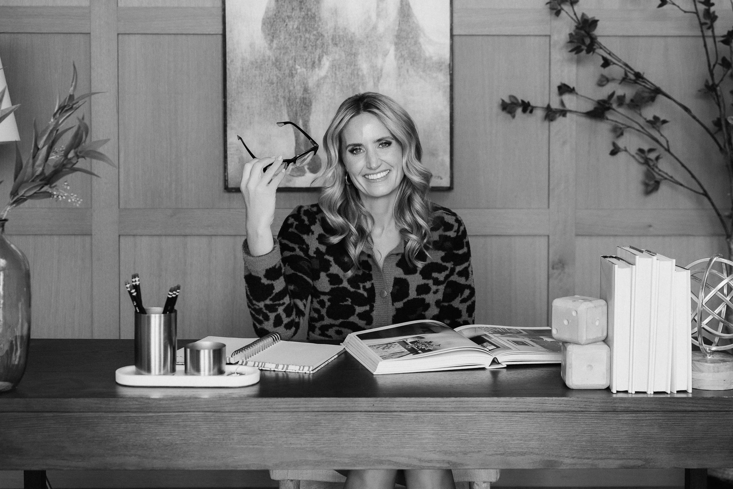 A woman with wavy hair smiling, holding glasses, sitting at a desk with books, notebook, pens, and decorative items, in a modern office with wood paneling and plants.
