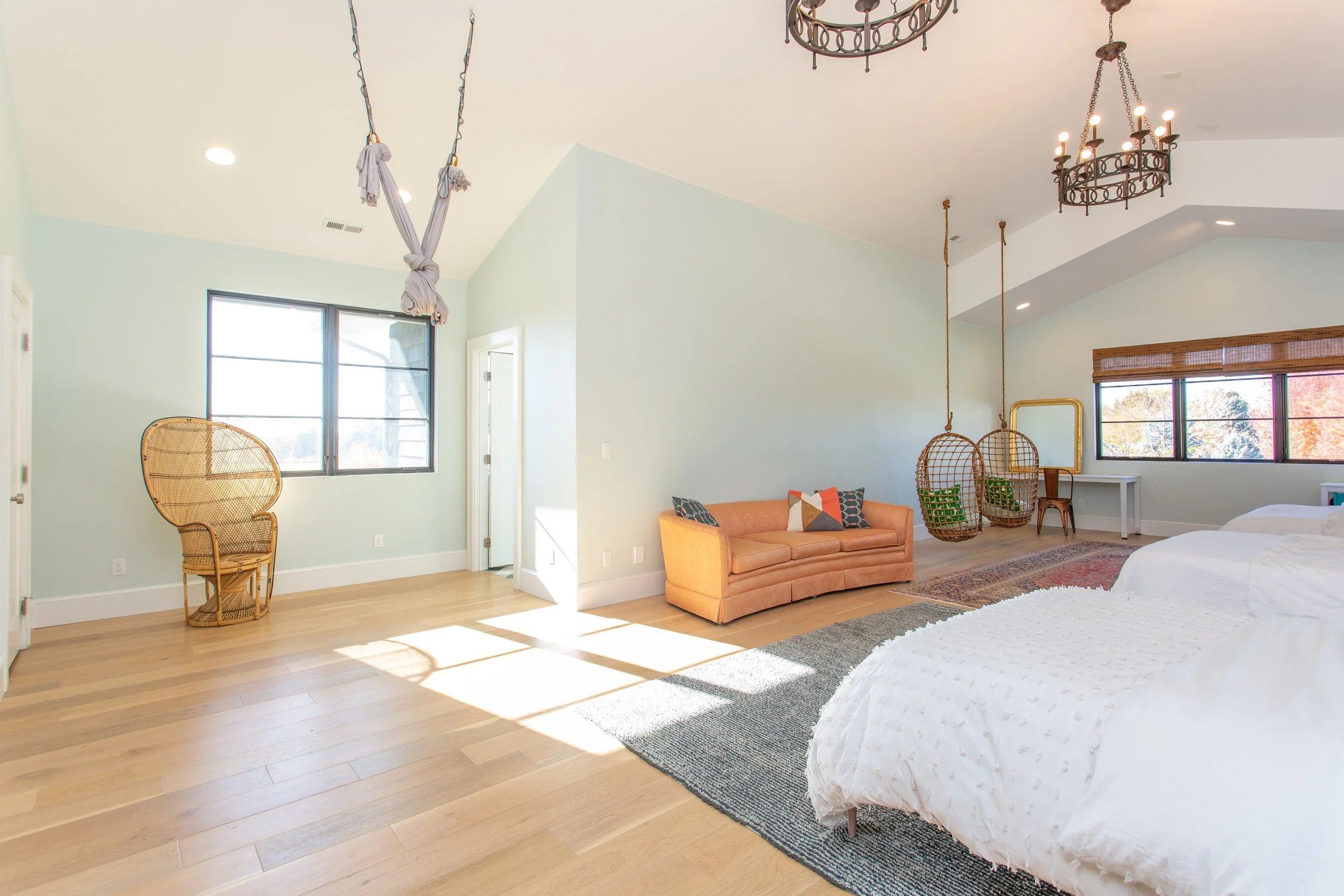 A spacious, well-lit bedroom with light wood flooring, featuring two beds, a pink sofa with pillows, two hanging wicker chairs, a white desk, an ornate chandelier, and large windows with black frames and brown blinds.