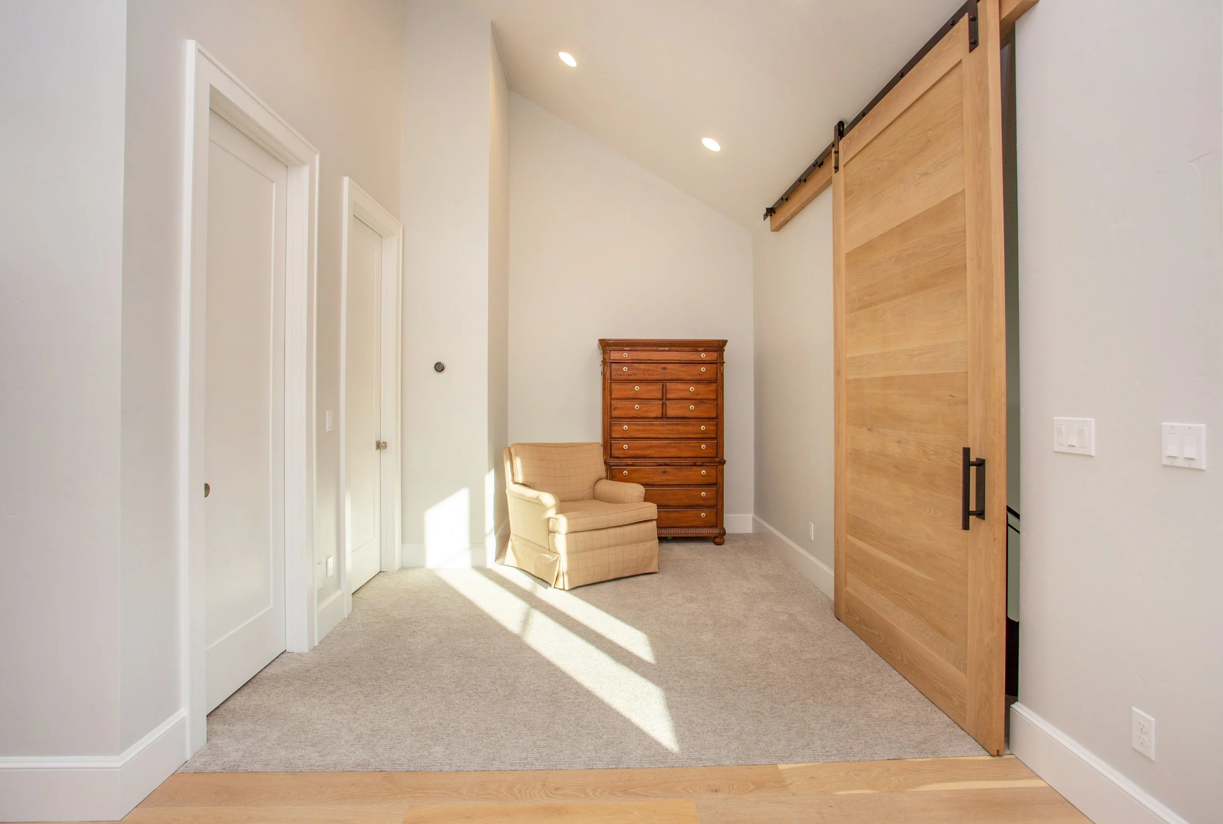 Room with beige carpet, white walls, a wooden sliding barn door, a small upholstered armchair, a wooden chest of drawers, and sunlight streaming in.