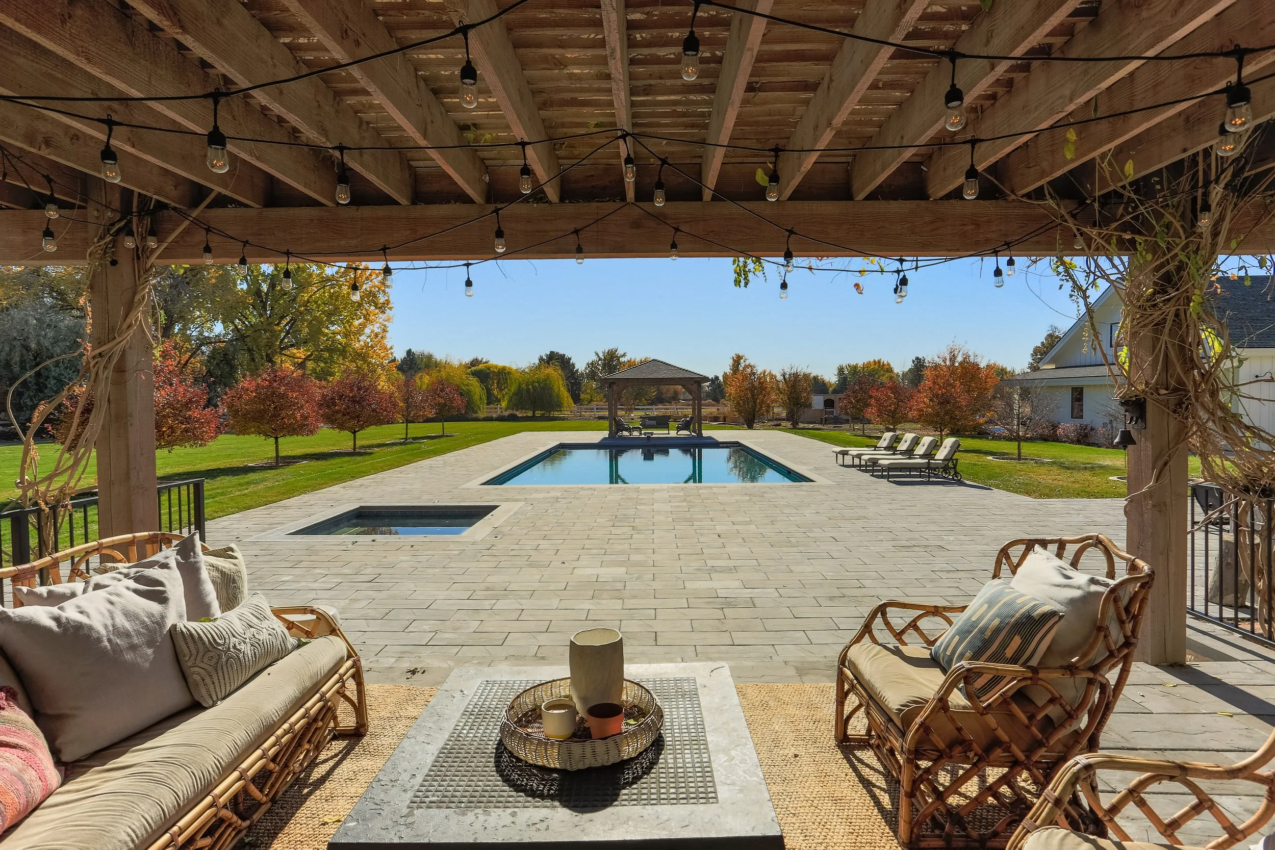 A backyard patio with outdoor seating, a swimming pool, trees with fall foliage, and a gazebo in the distance viewed from under a wooden pergola with string lights.