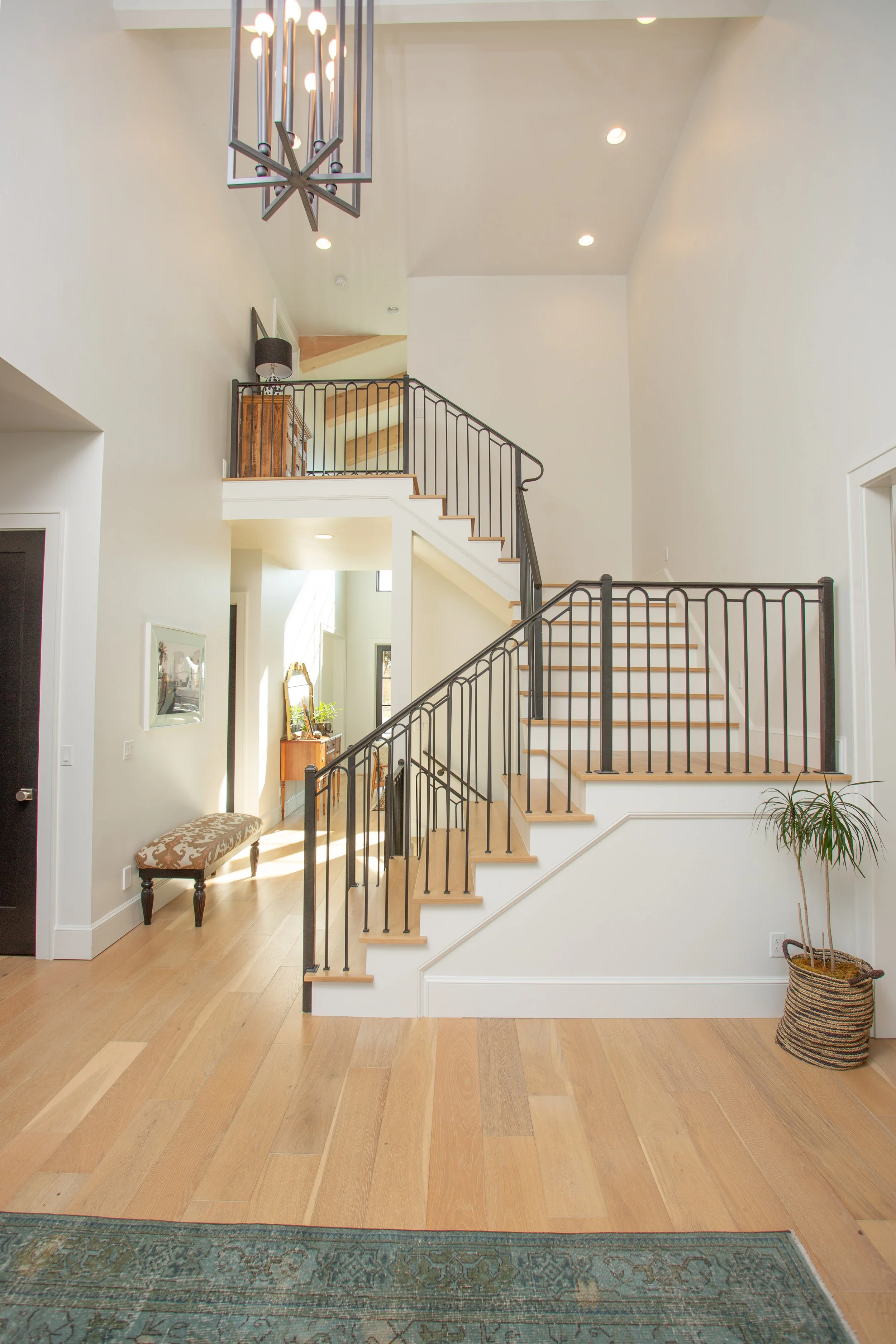 Interior view of a modern home with a staircase featuring black metal railings, a wooden banister, and white walls. There is a chandelier hanging from the ceiling, a decorative bench on the left, and a potted plant on the right. The hardwood floor ex