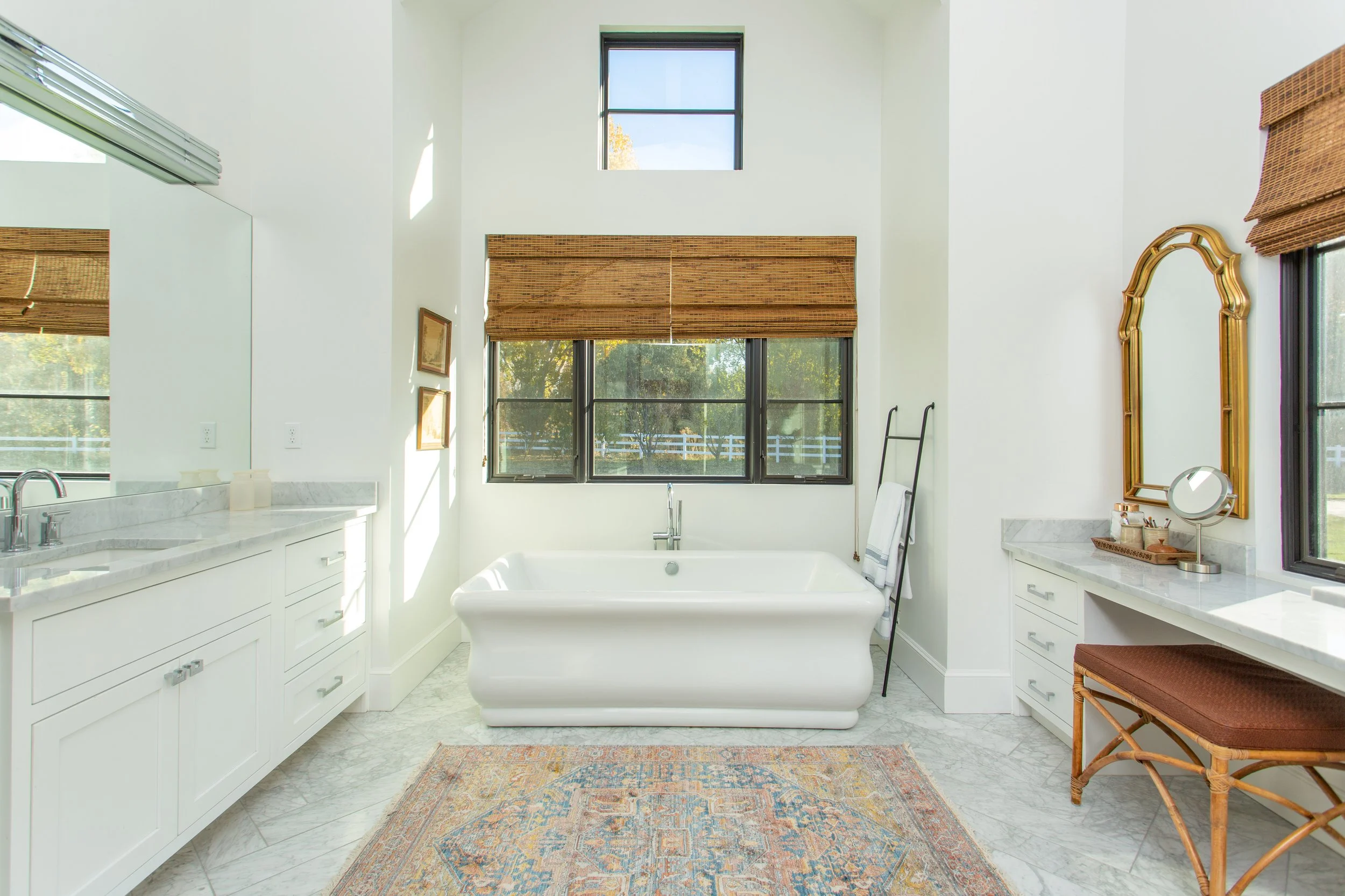 Bright bathroom with a freestanding bathtub, large windows with bamboo blinds, white cabinetry, marble countertops, a mirror, and a patterned rug.