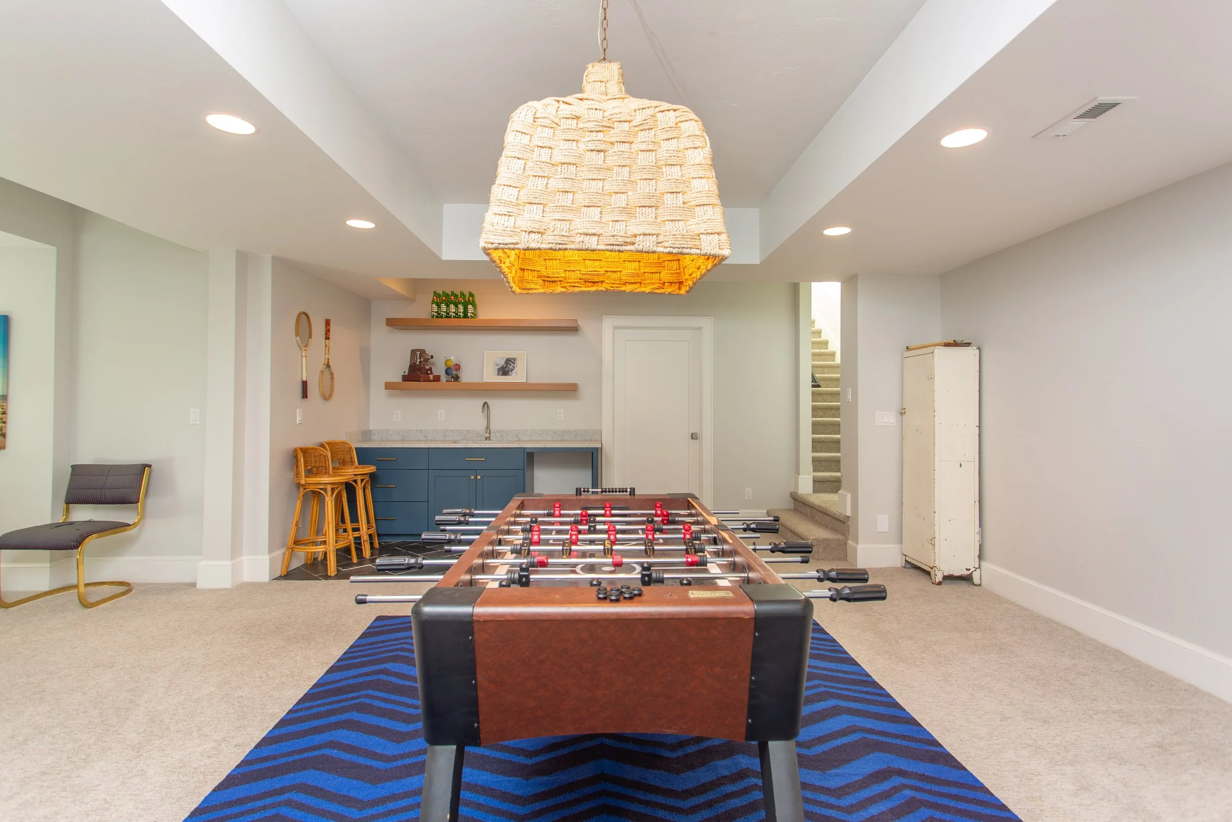 Indoor recreation room with foosball table on blue striped rug, wicker barstools, blue kitchenette, and staircase in background.
