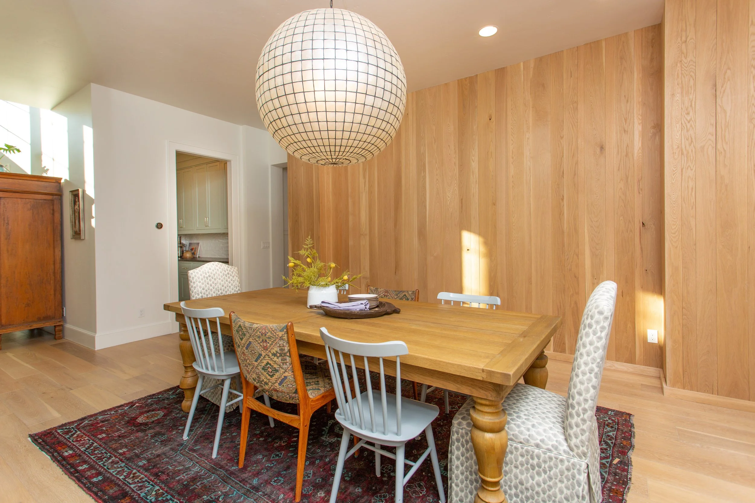 Dining room with a large wooden table, mixed chairs, a patterned rug, wood-paneled wall, and a spherical hanging light fixture.
