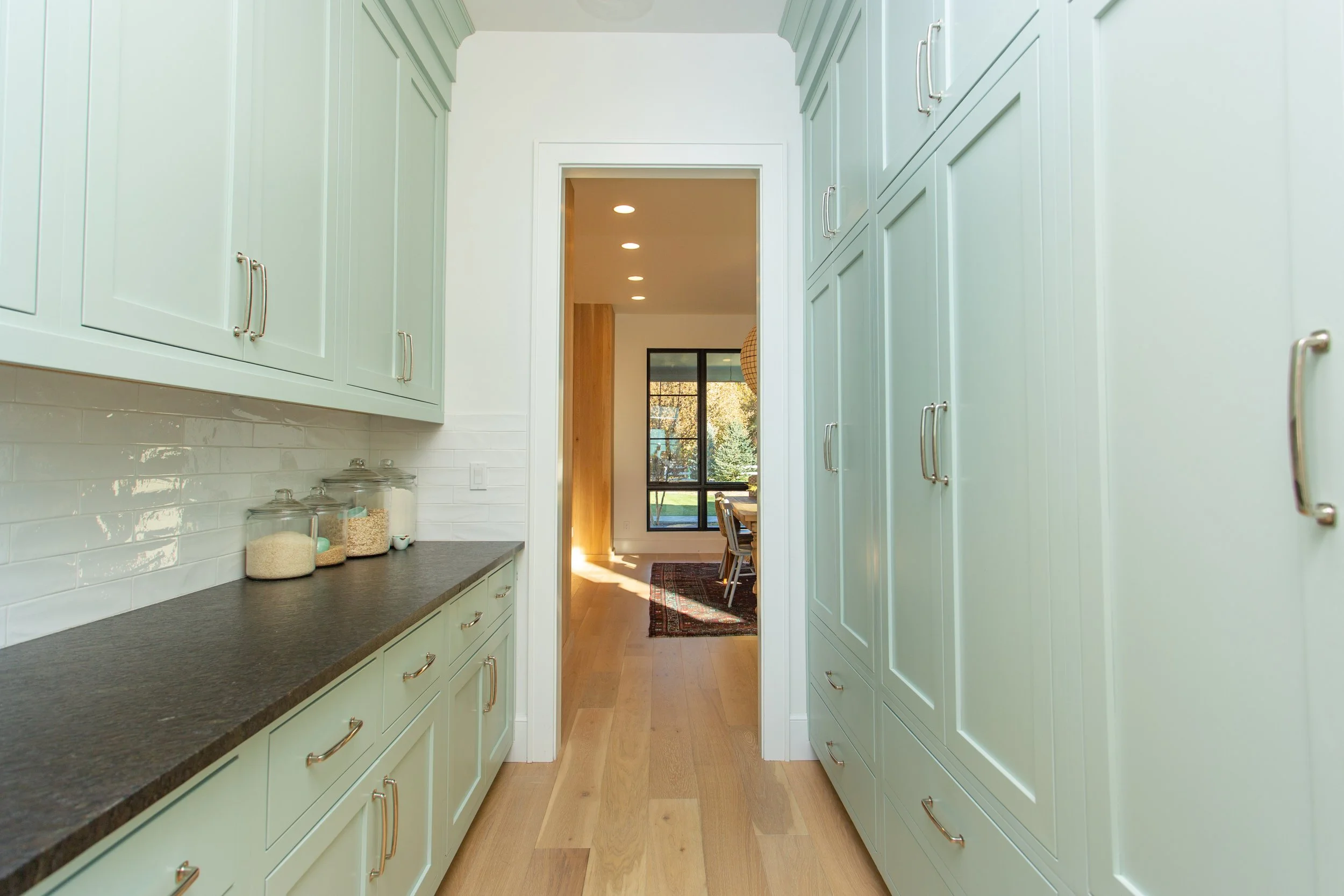 Kitchen galley with light green cabinets, dark countertop, visible pantry with storage jars, leading to a dining area with large windows and wooden flooring.