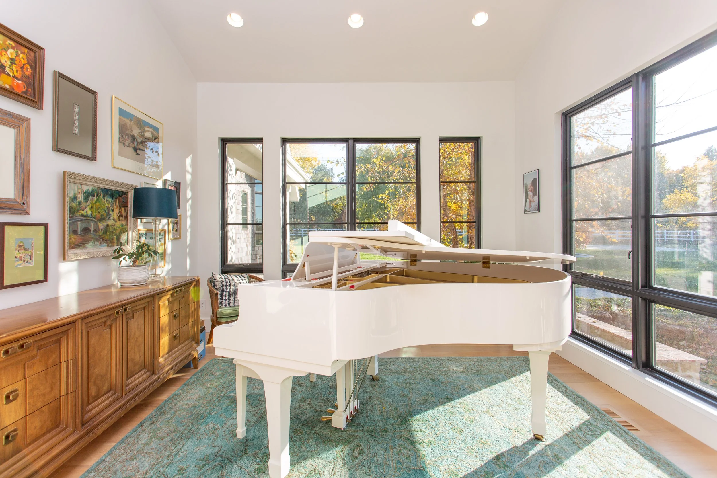 A white grand piano in a sunlit room with large windows, a wooden sideboard decorated with framed artwork, a potted plant, and a chair with a cushion.