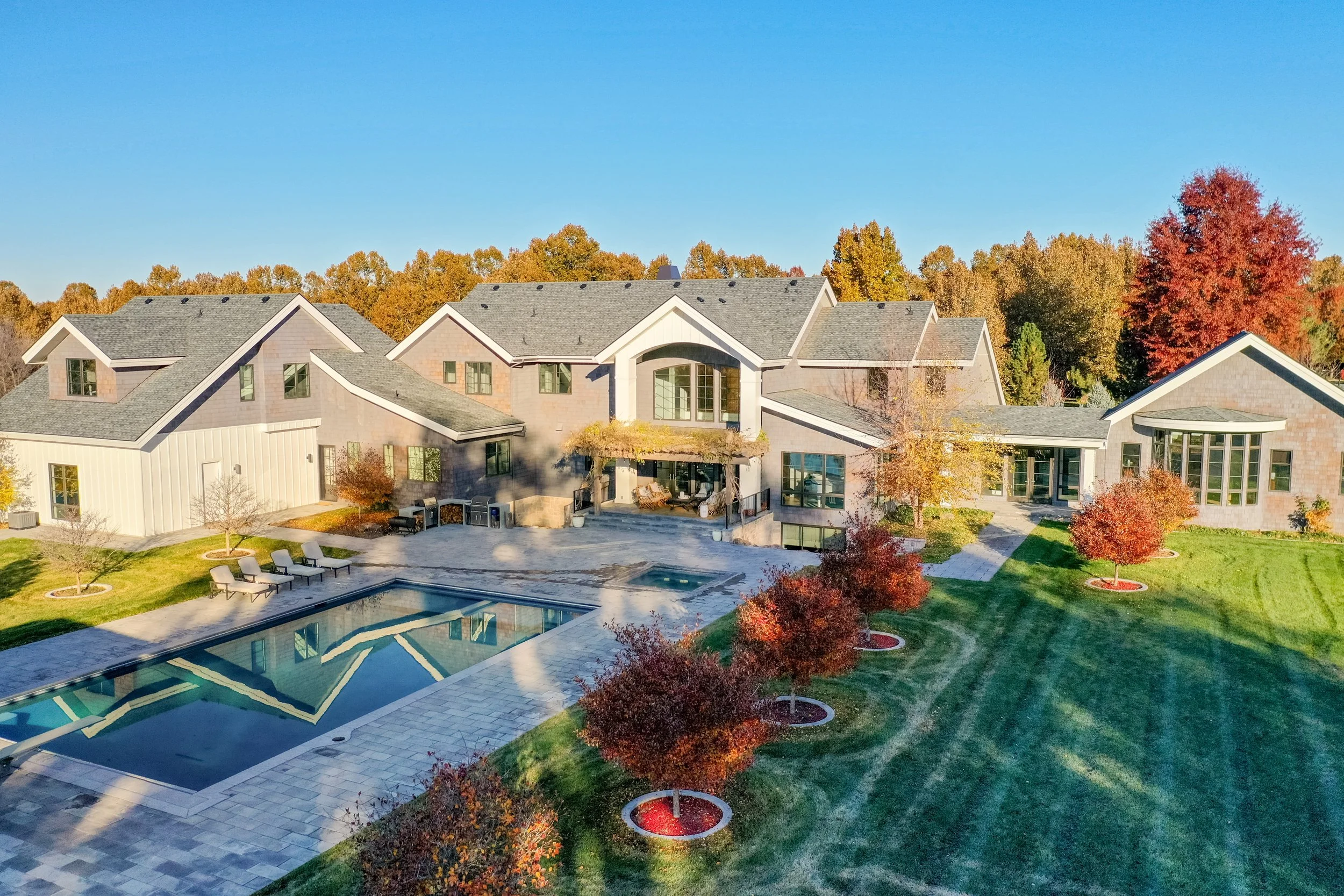 A large modern house with gray and white exterior, situated in a landscaped yard with a swimming pool, lounge chairs, and red-leafed trees, surrounded by fall-colored foliage.