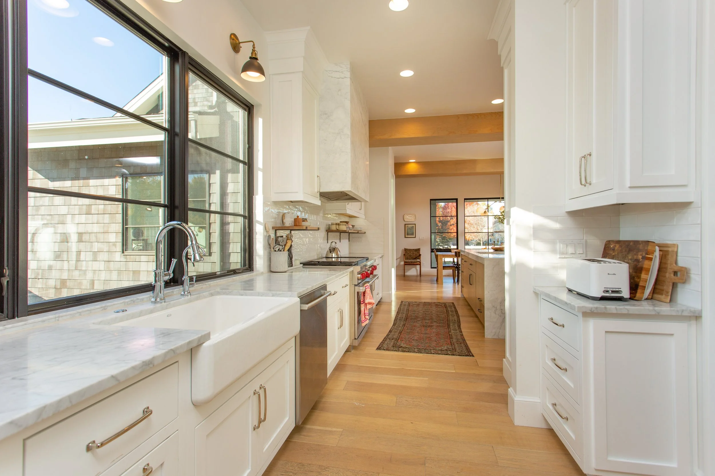 Bright modern kitchen with large window, white cabinets, marble countertops, stainless steel appliances, and hardwood floors, with dining area visible in the background.