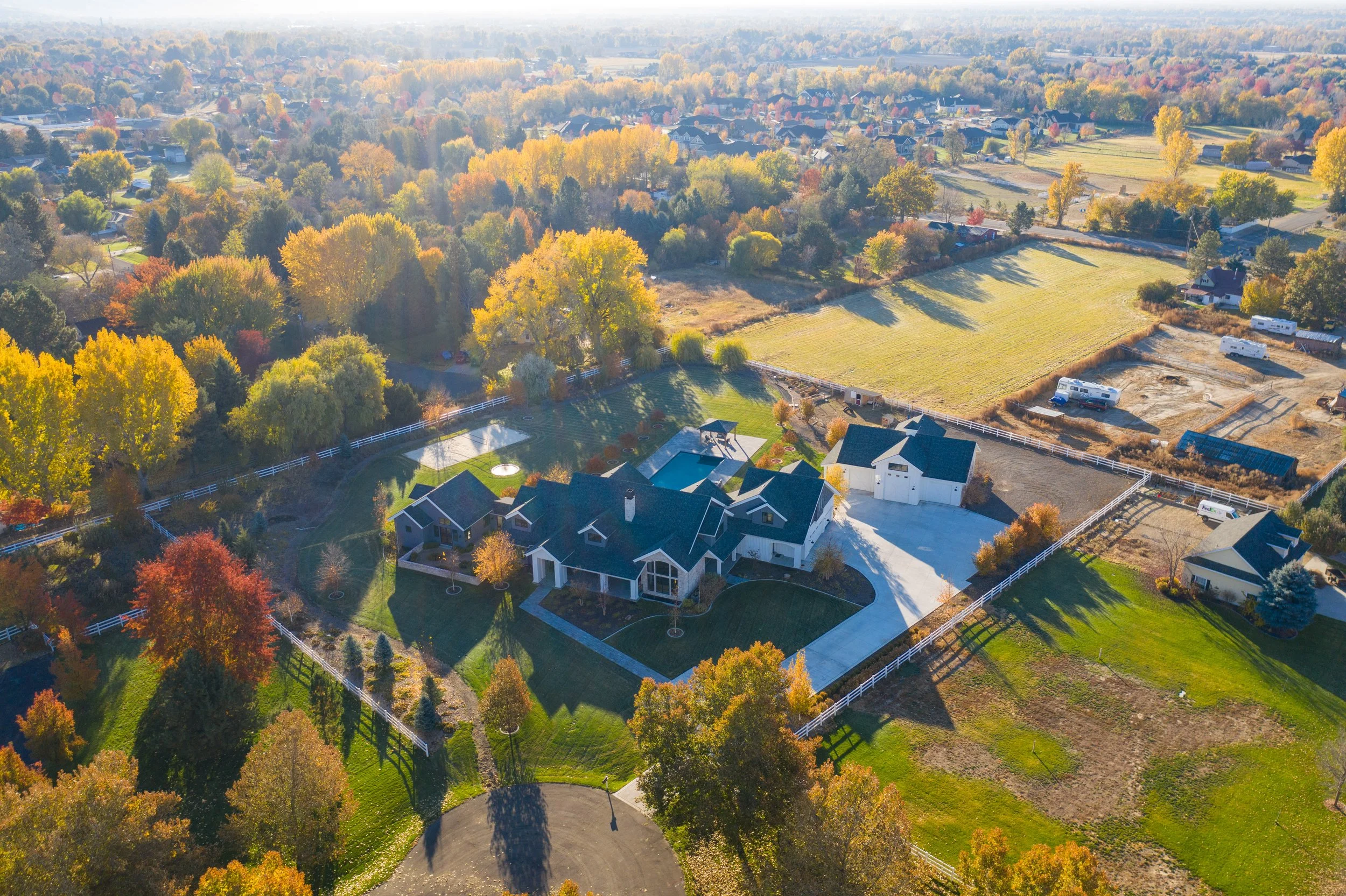 Aerial view of a large house with a black roof, swimming pool, and expansive green lawn, surrounded by trees with autumn foliage, white fence, and neighboring houses and open fields.