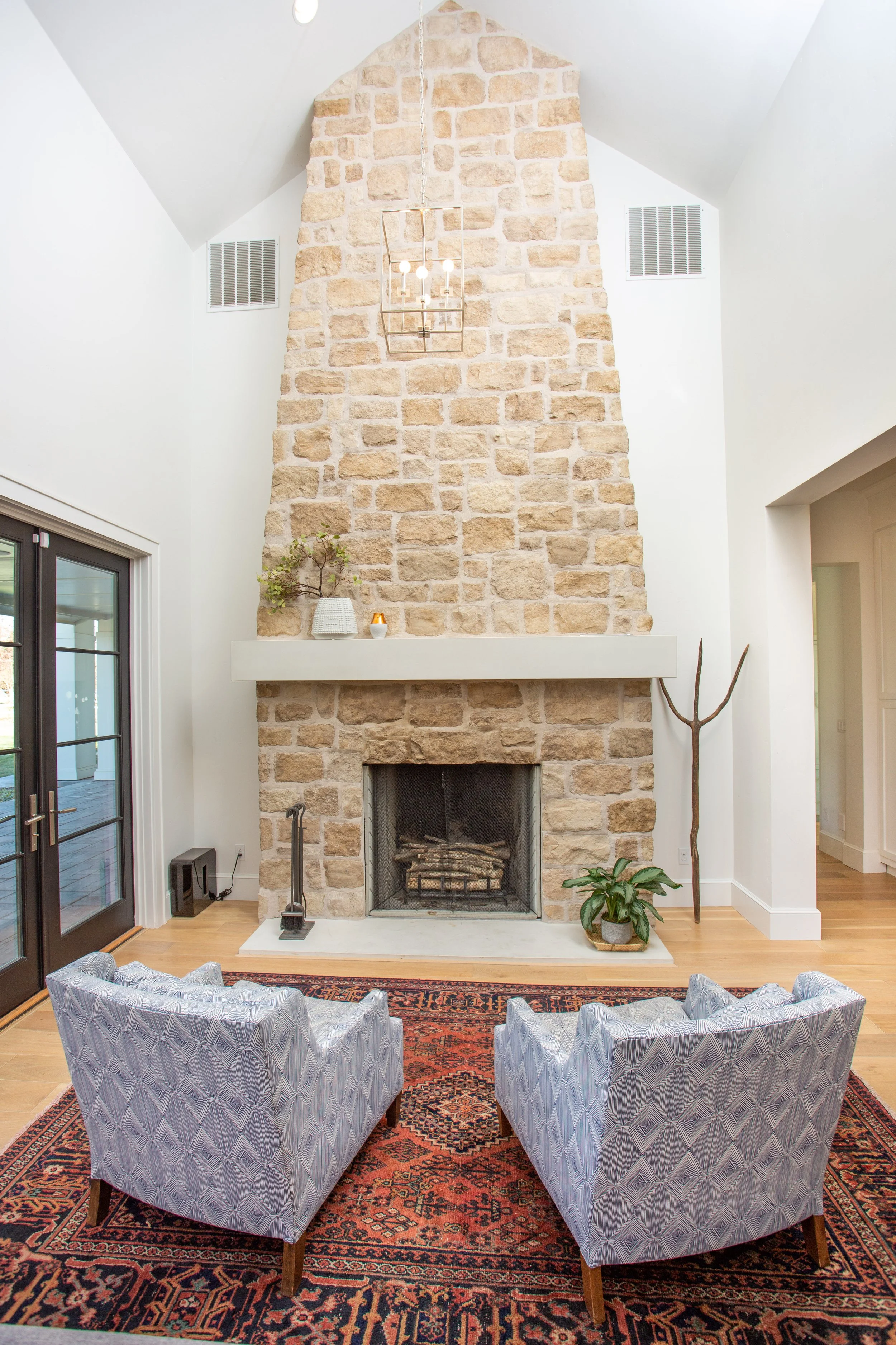 Living room with a tall stone fireplace, two patterned armchairs on a red and black rug, a plant and decorative branches near the fireplace, and a glass sliding door.