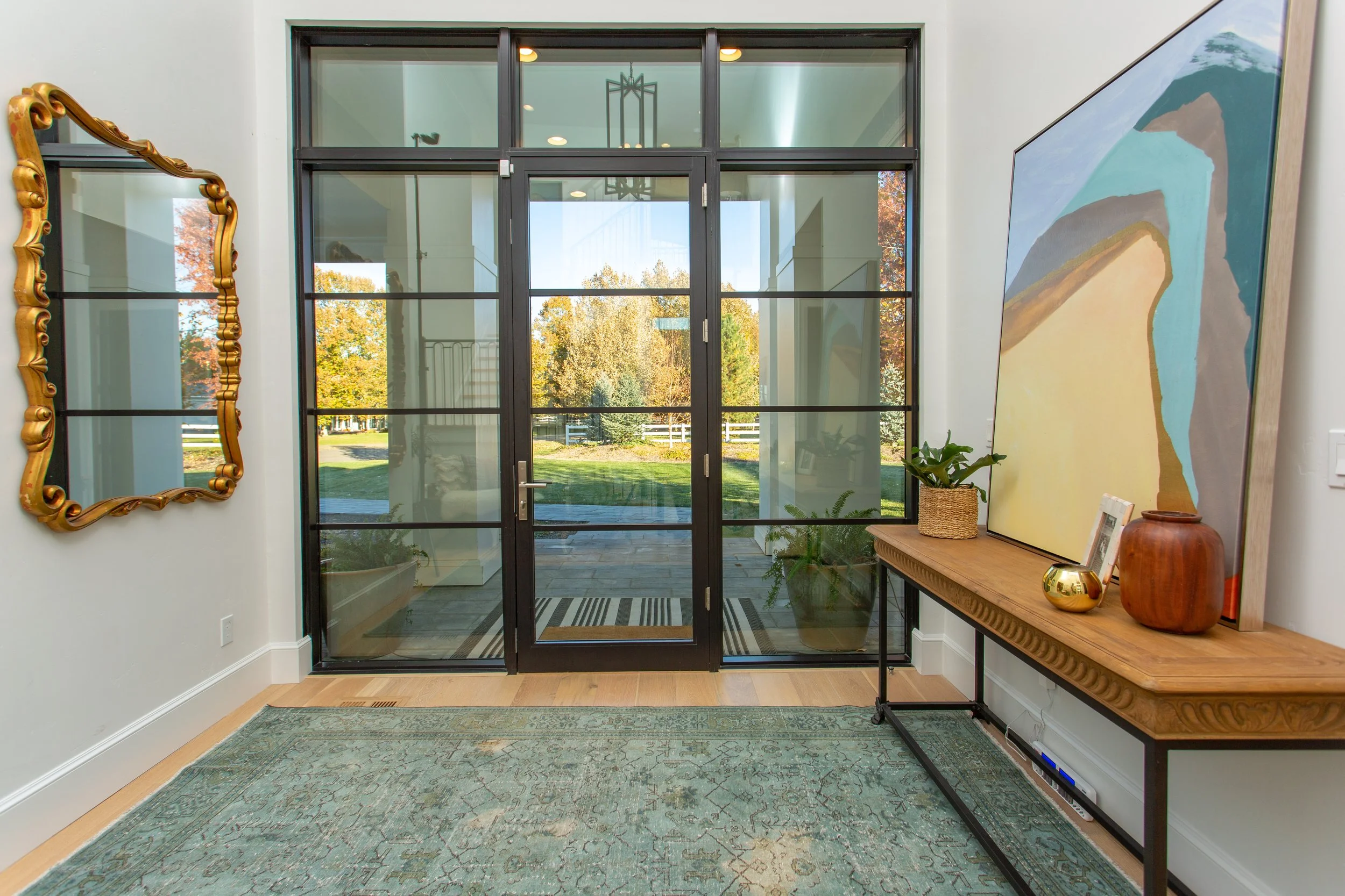 Bright entryway with a large glass door and windows, decorative gold mirror on the left wall, a table with plants and artwork on the right, and an outdoor view of trees through the glass.