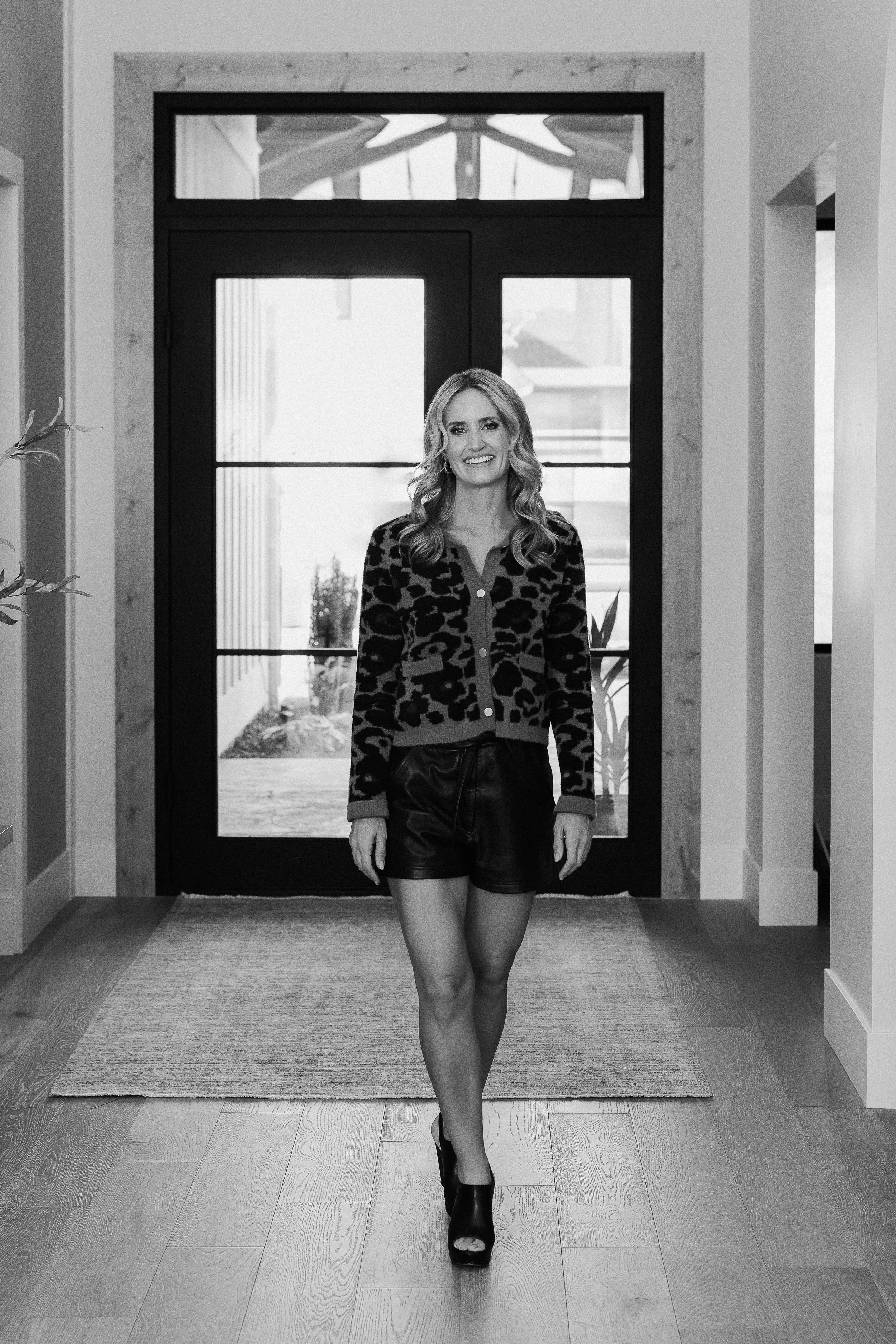 A woman with wavy hair smiling, standing in a hallway near a front door, wearing a leopard print cardigan, leather shorts, and high heels.