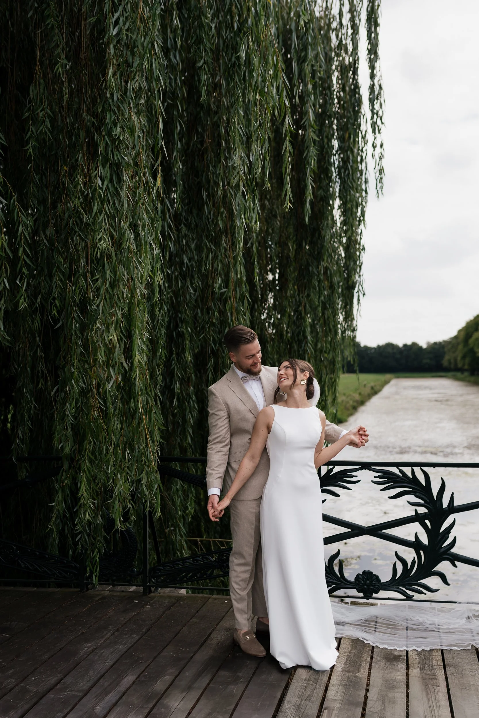 Ein Paar in Hochzeitskleidung, das sich liebevoll auf einer Brücke an einem Fluss ansieht, mit üppigem Grün und einem bewölkten Himmel im Hintergrund.
