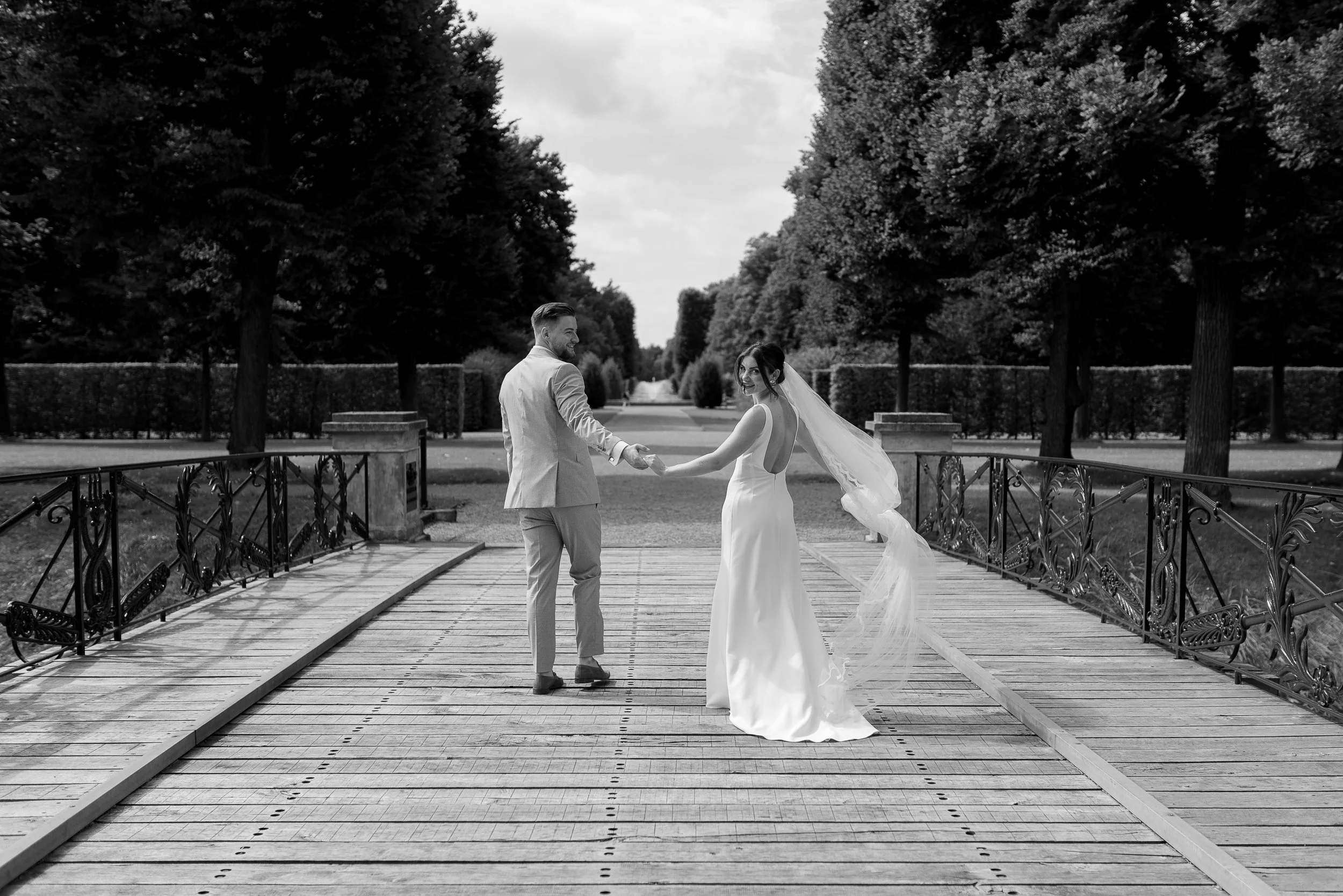 Ein Brautpaar hält sich an einem Baumarkt, während sie auf einer Brücke in einem Park stehen. Es ist schwarz-weiß Bilder, das Paar trägt Hochzeitskleidung, die Braut trägt ein langes Kleid mit Schleier, der Bräutigam trägt einen Anzug. Im Hintergrund