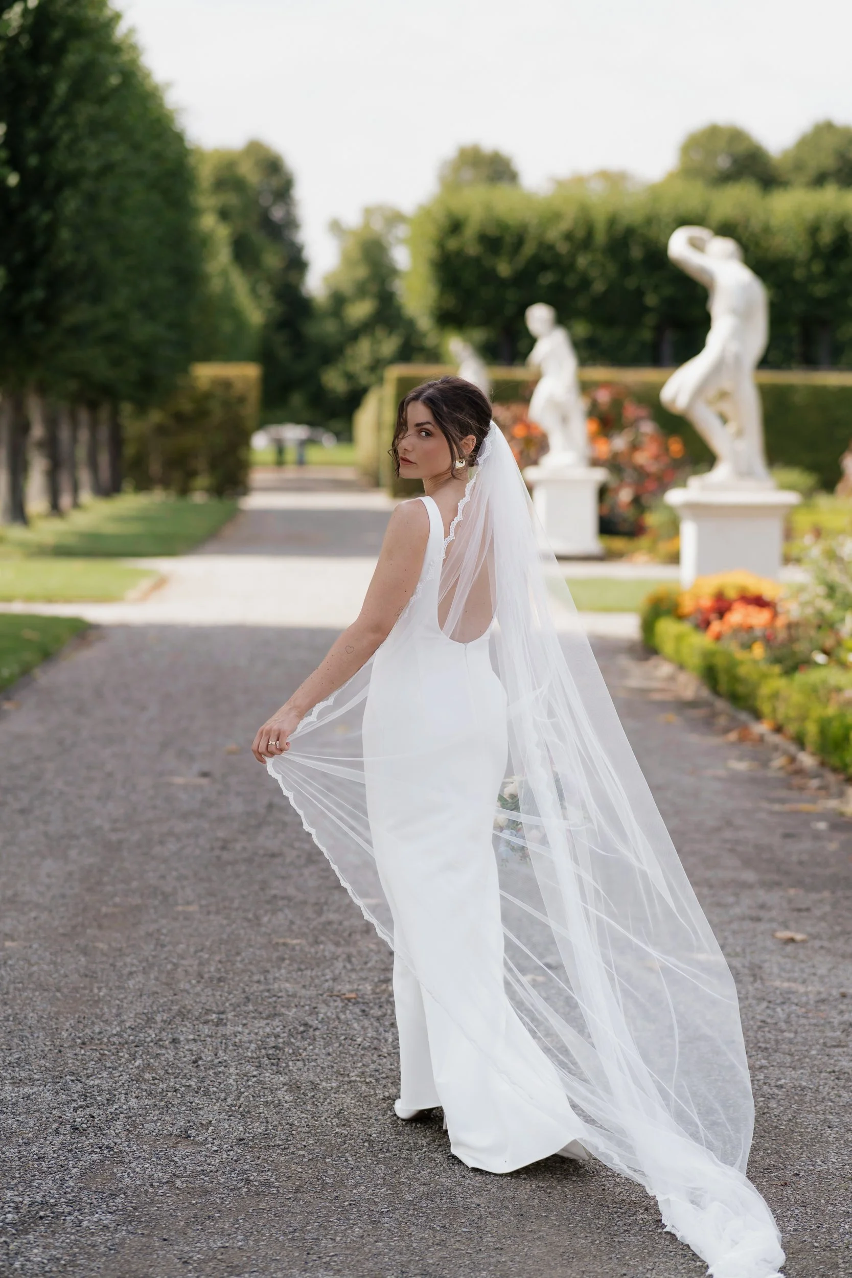 Eine junge Frau im weißen Brautkleid mit Schleier steht auf einem gesteinerten Weg in einem Garten, im Hintergrund sind Skulpturen und grüne Bäume zu sehen.