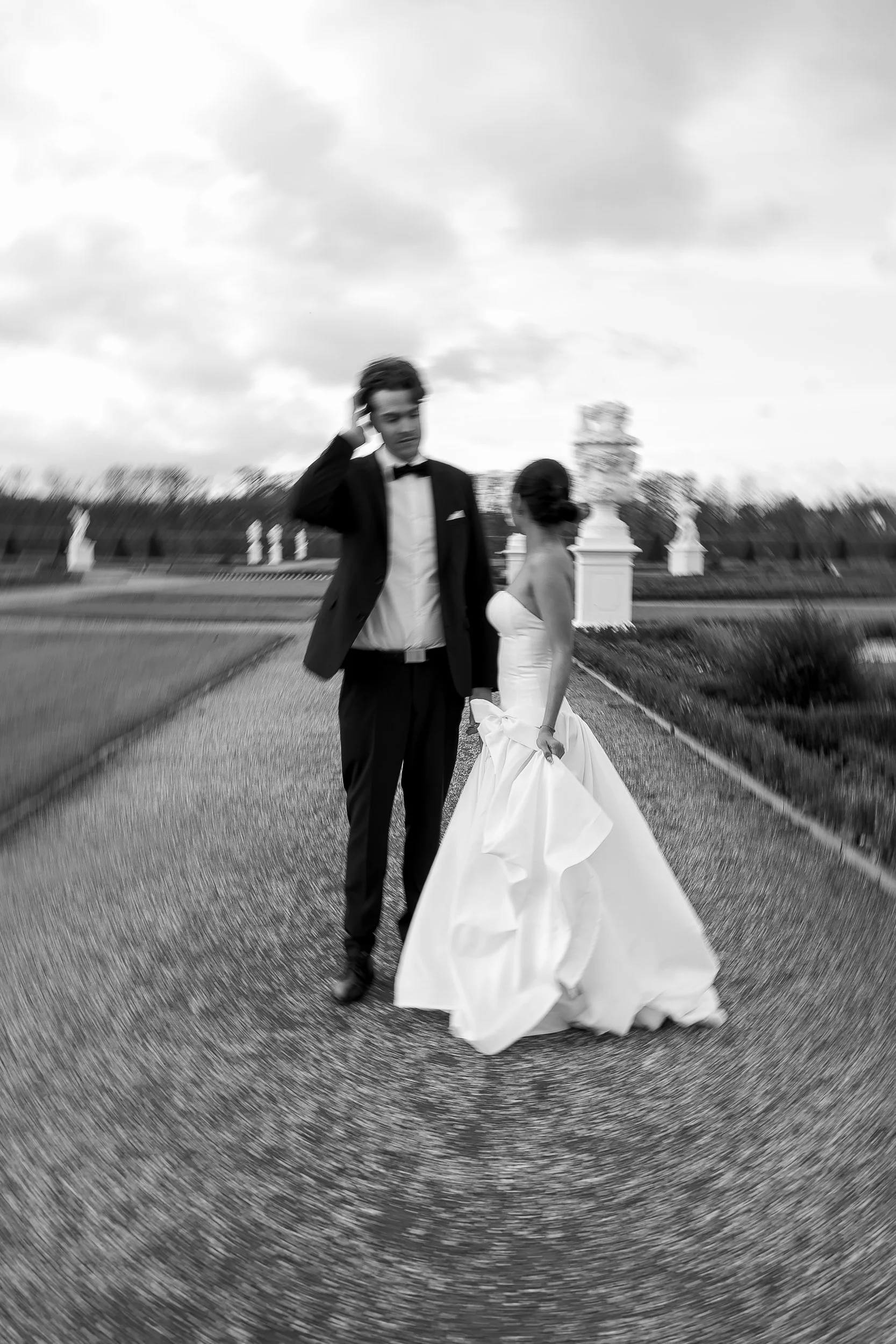 Schwarz-weiß Foto eines Paares in formeller Kleidung, vermutlich Hochzeitskleidung, bei einem Spaziergang im Freien, umgeben von Statuen und Bäumen, mit einem Wolkenhimmel im Hintergrund
