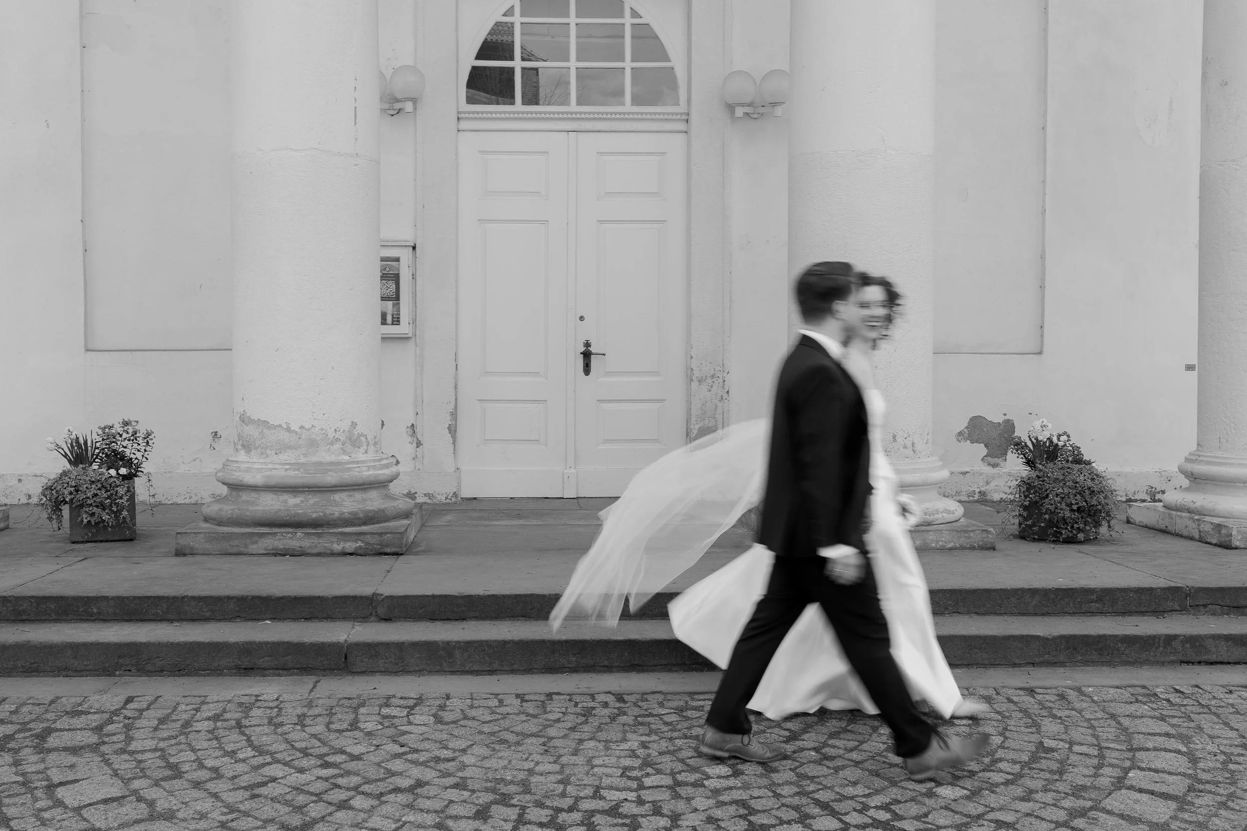 Zwei Menschen in formeller Kleidung, einer trägt einen Hochzeitsanzug und das andere einen weißen Brautkleid, gehen nebeneinander an einer weiß gestrichenen Gebäudefront mit großen Säulen vorbei.