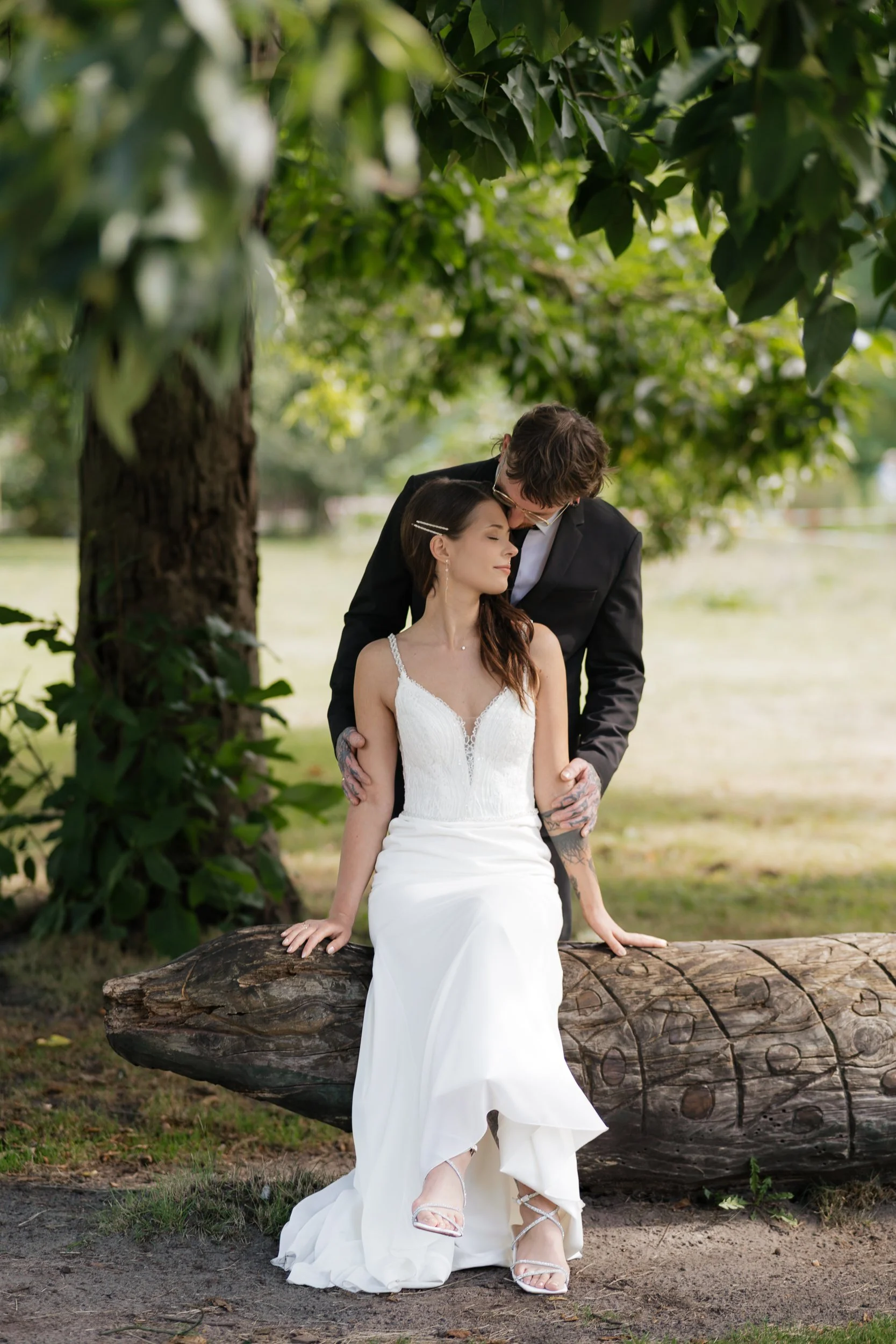 Ein Brautpaar sitzt auf einem Baumstamm in einem Park, umgeben von Bäumen und Grünflächen, bei einem Fotoshooting am Tag.