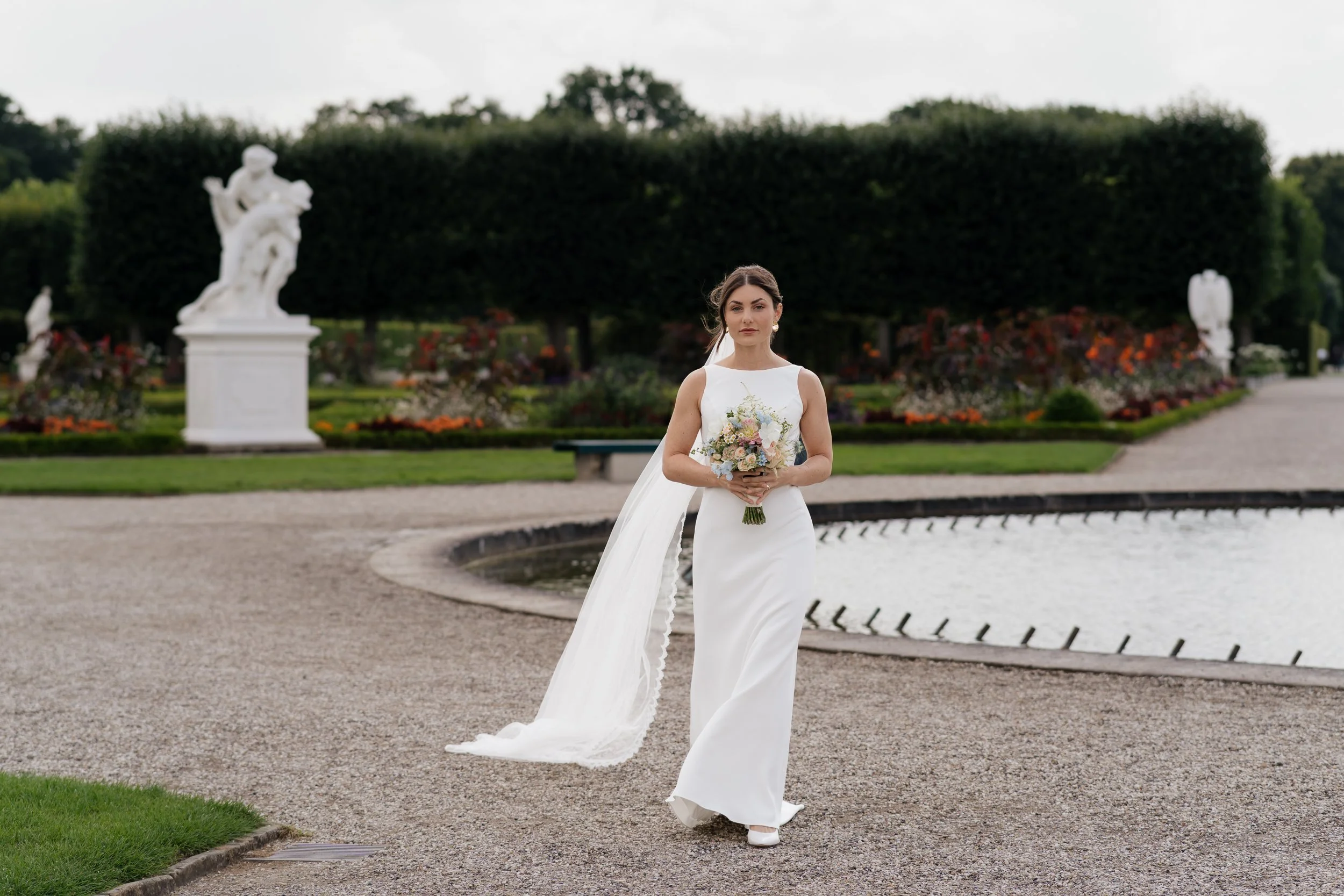 Bräutigam in weißem Hochzeitsanzug hält Blumenstrauß im Garten mit Brunnen und Skulpturen im Hintergrund.