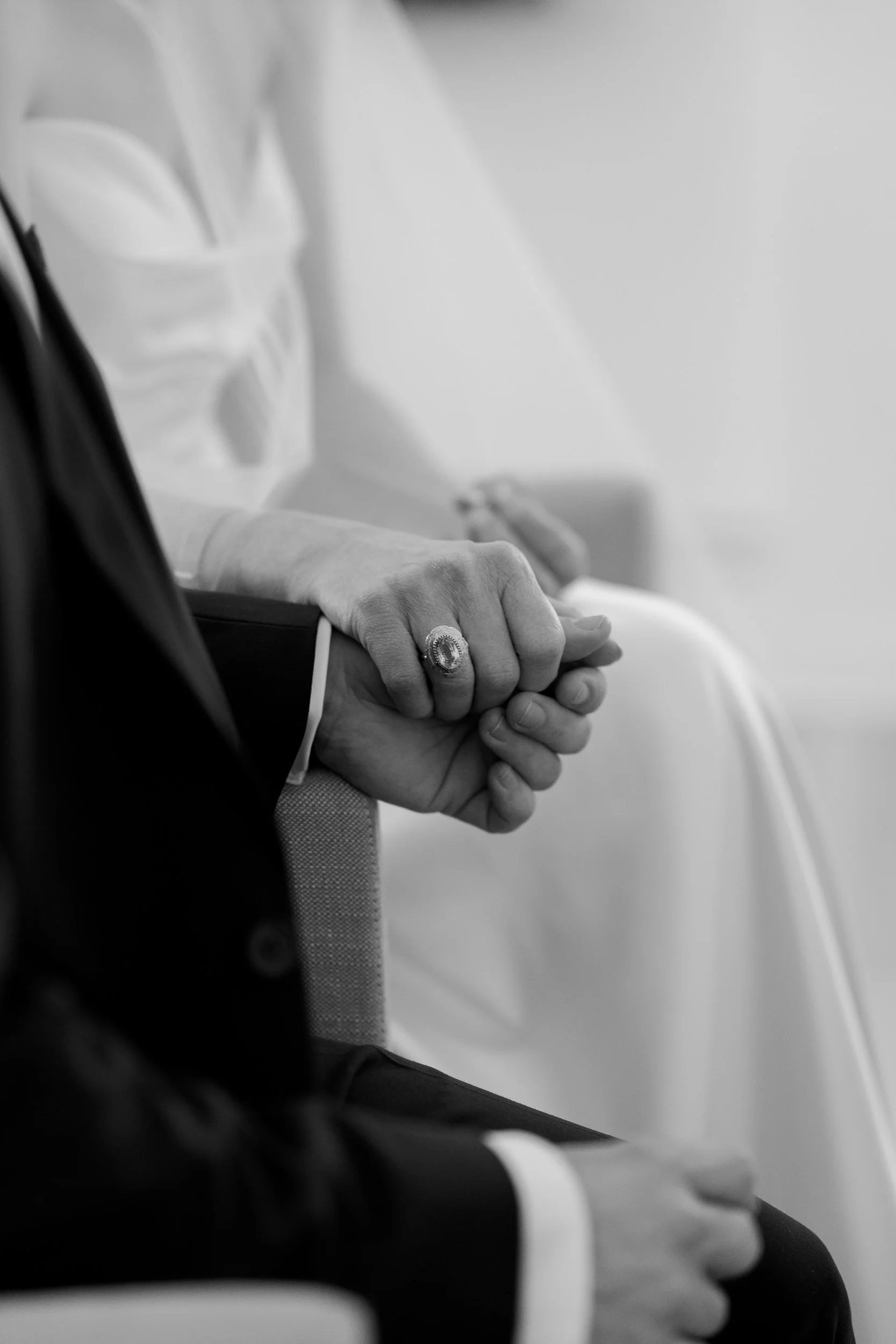 Nahaufnahme von zwei Personen, die Händchen halten, mit Fokus auf die Hände. Eine Person trägt einen großen Ring. Das Bild ist in Schwarz-Weiß. Der Hintergrund ist unscharf.