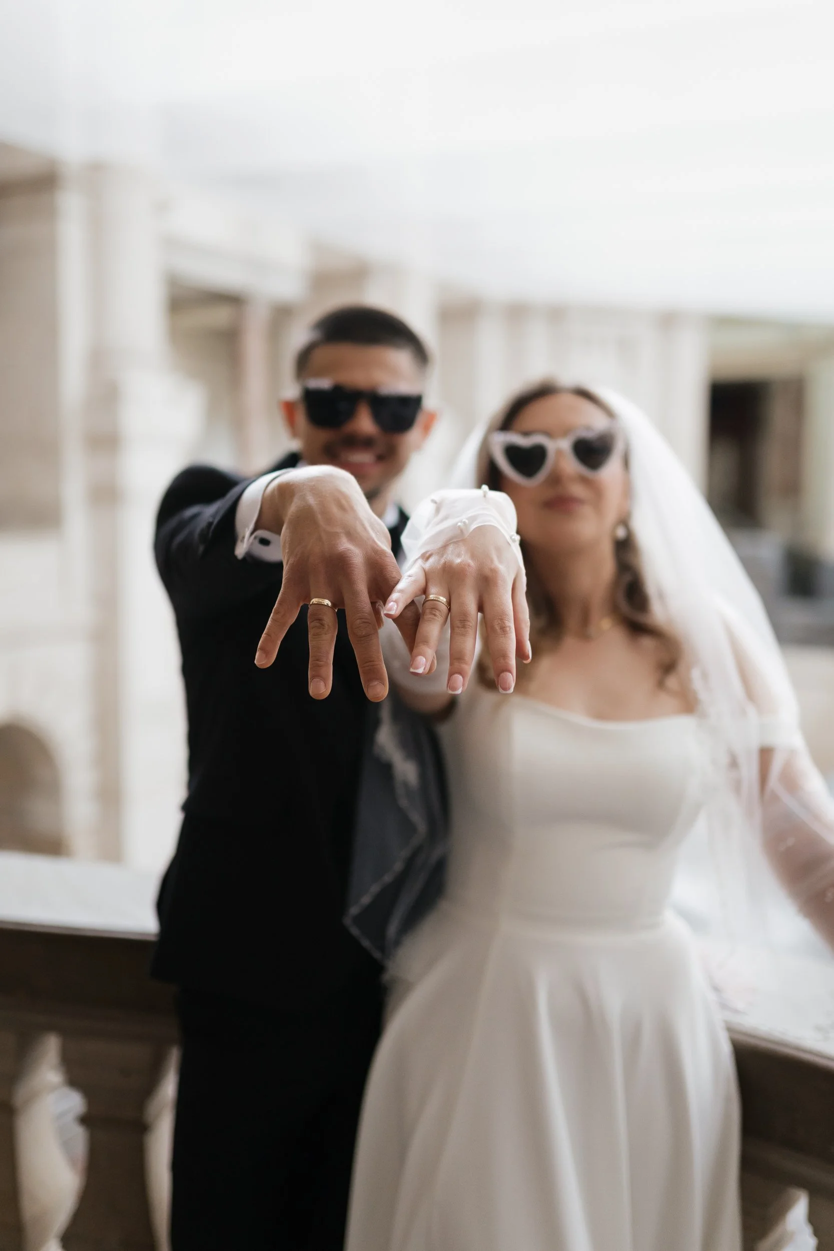 Ein Brautpaar bei einer Hochzeit zeigt ihre Eheringe, beide tragen Sonnenbrillen und lächeln.