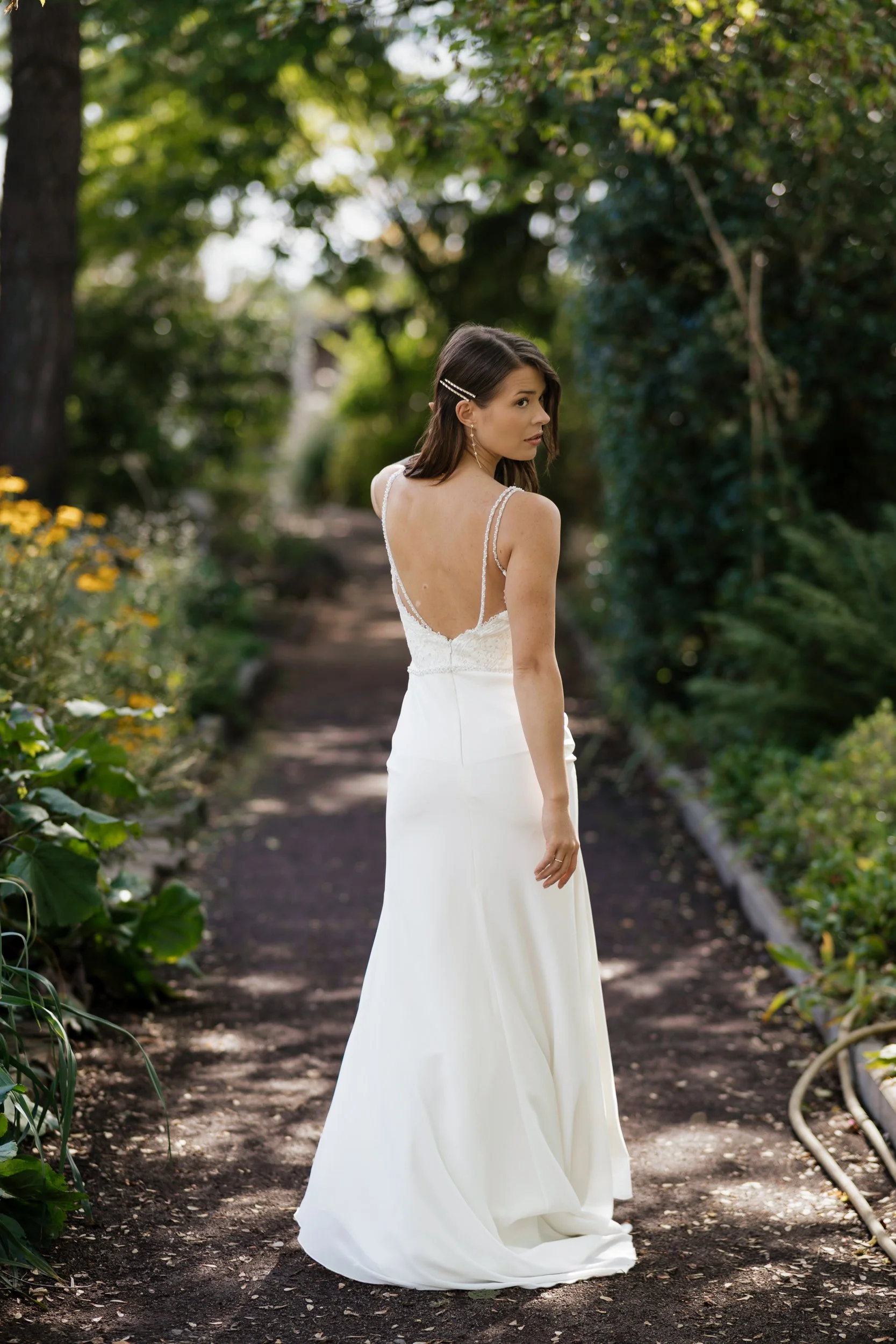 Eine Frau trägt ein weißes Brautkleid und steht auf einem Waldweg, umgeben von grünen Bäumen und Blumen.