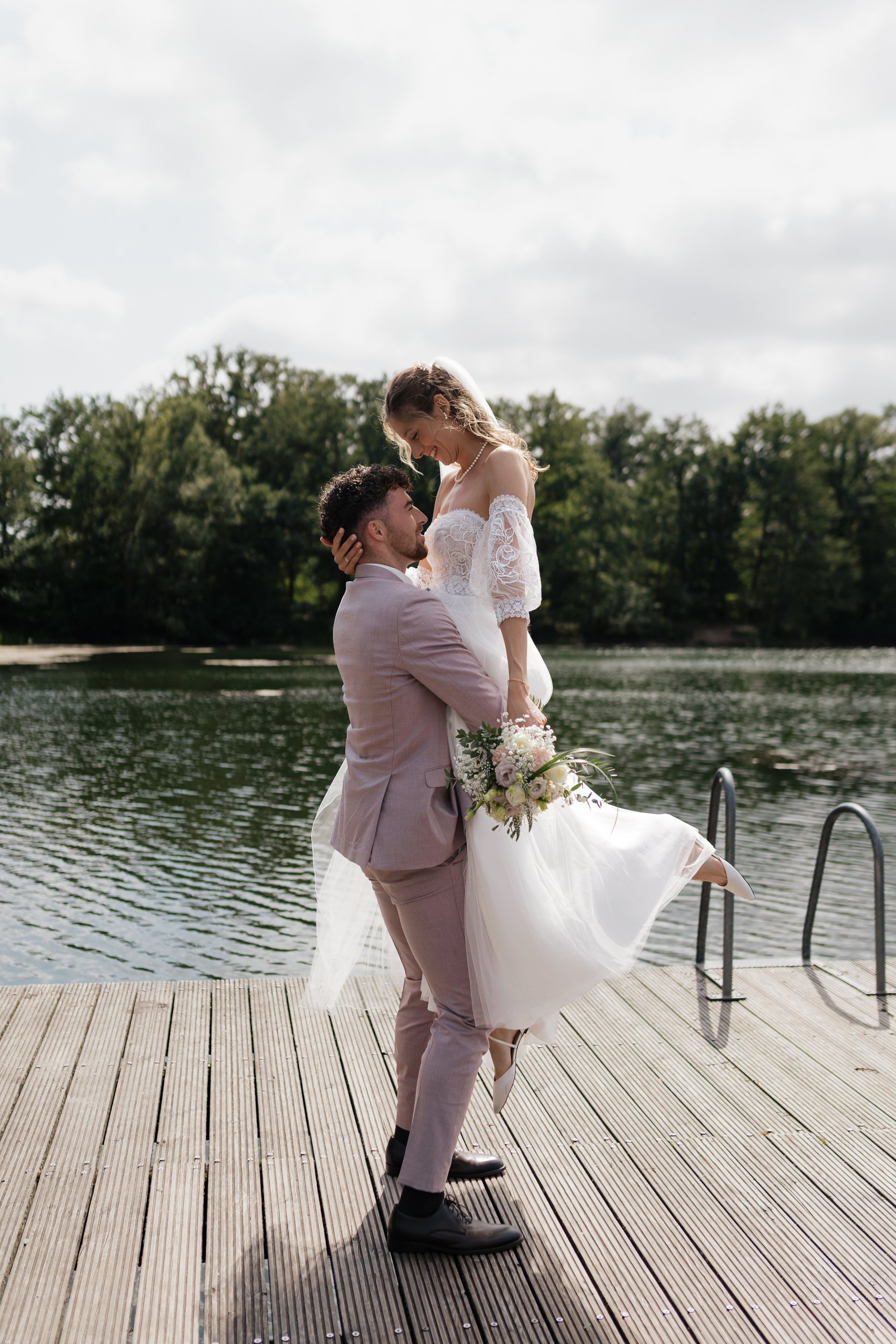 Ein Brautpaar am Wasser, der Bräutigam trägt einen hellen Anzug, die Braut in einem weißen Kleid hält einen Blumenstrauß, sie lachen und umarmen sich auf einem Holzsteg mit einem Wasser im Hintergrund und einem bewölkten Himmel.