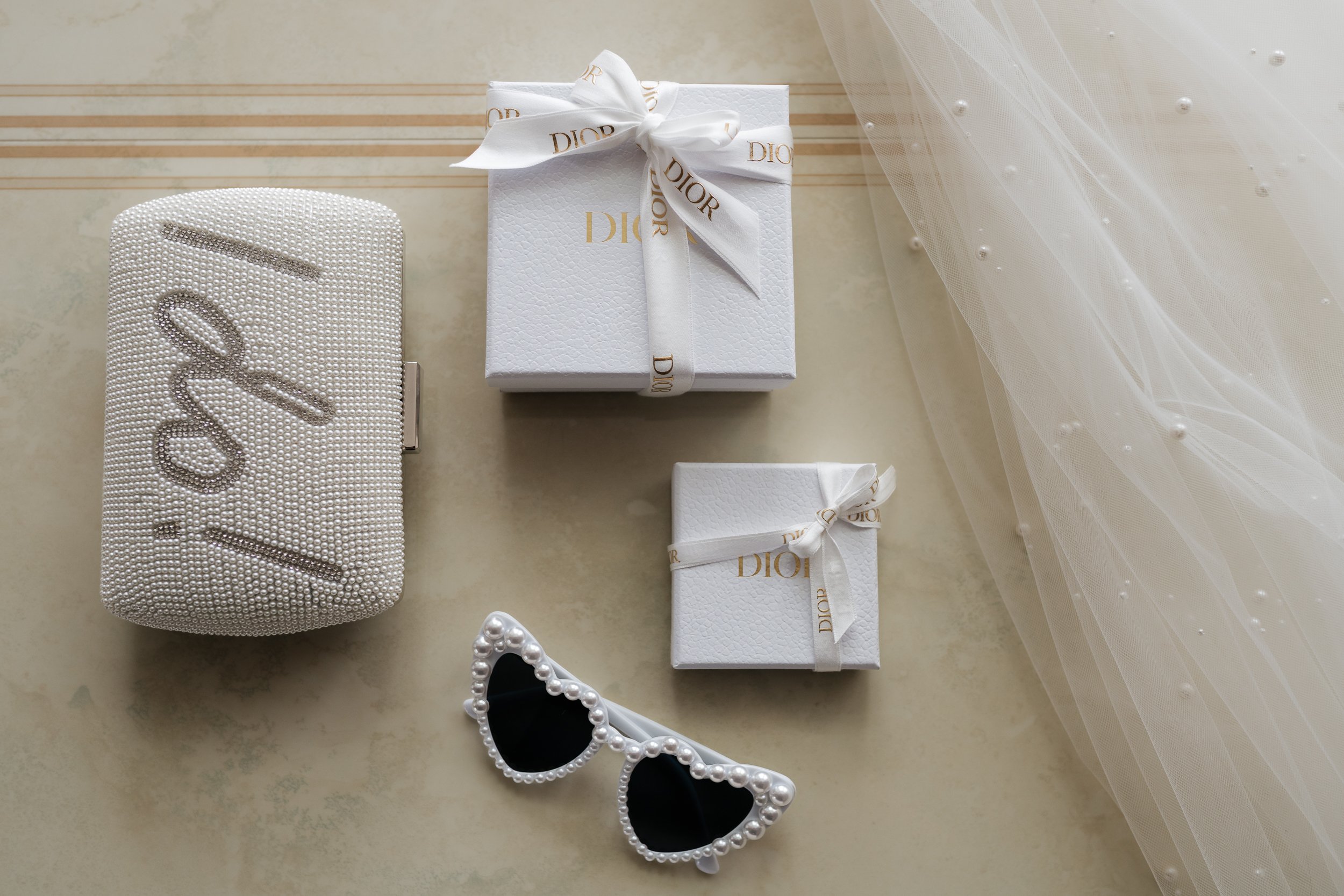 Weißes Geschenk mit goldener Beschriftung 'Dior' und goldener Schleife, eine kleine weiße Geschenkbox mit Schleife, eine weiße Handtasche mit Perlen-Design, Sonnenbrille mit Perlenrahmen, schwarze Gläser, Eiskristall-Haarreifen, Tüllstoff mit Perlen